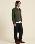 A man stands in profile wearing an olive green jacket over a brown knit sweater, white collared shirt, black trousers, and brown leather shoes, against a plain warm beige studio background.