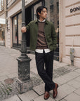 A man stands on a city sidewalk wearing an olive green jacket over a brown sweater and white shirt, paired with dark trousers and brown leather shoes, leaning against a decorative black lamppost.