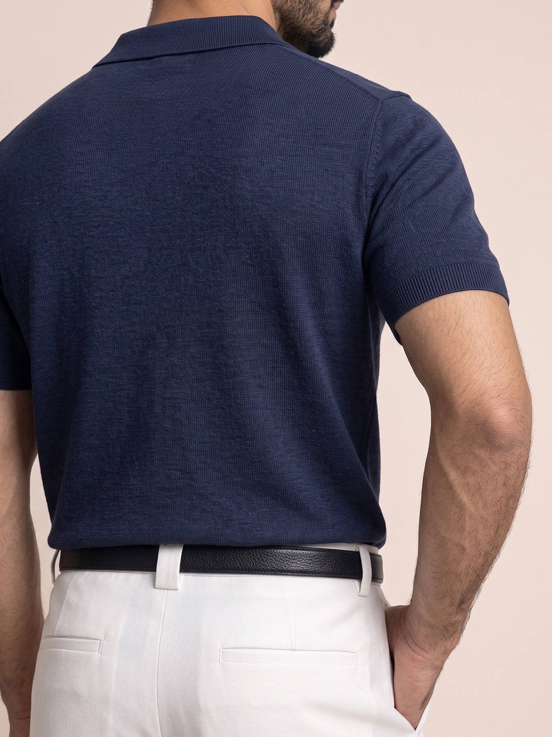 #blue-navy_group A man wearing a navy blue short-sleeve knit polo shirt, shown from the back. He is also wearing white trousers and a black belt, set against a solid pale peach background.