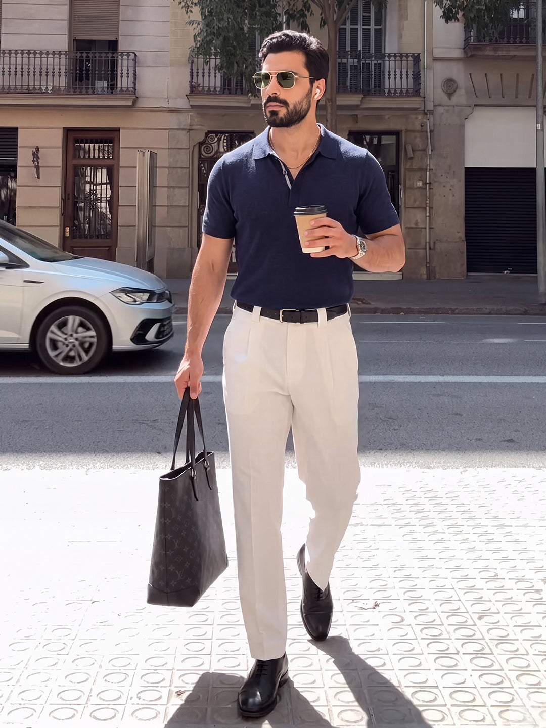 #blue-navy_group A man walking on a city sidewalk wearing a navy blue polo shirt, off-white trousers, black leather shoes, sunglasses, and carrying a black monogram tote bag and a coffee cup.