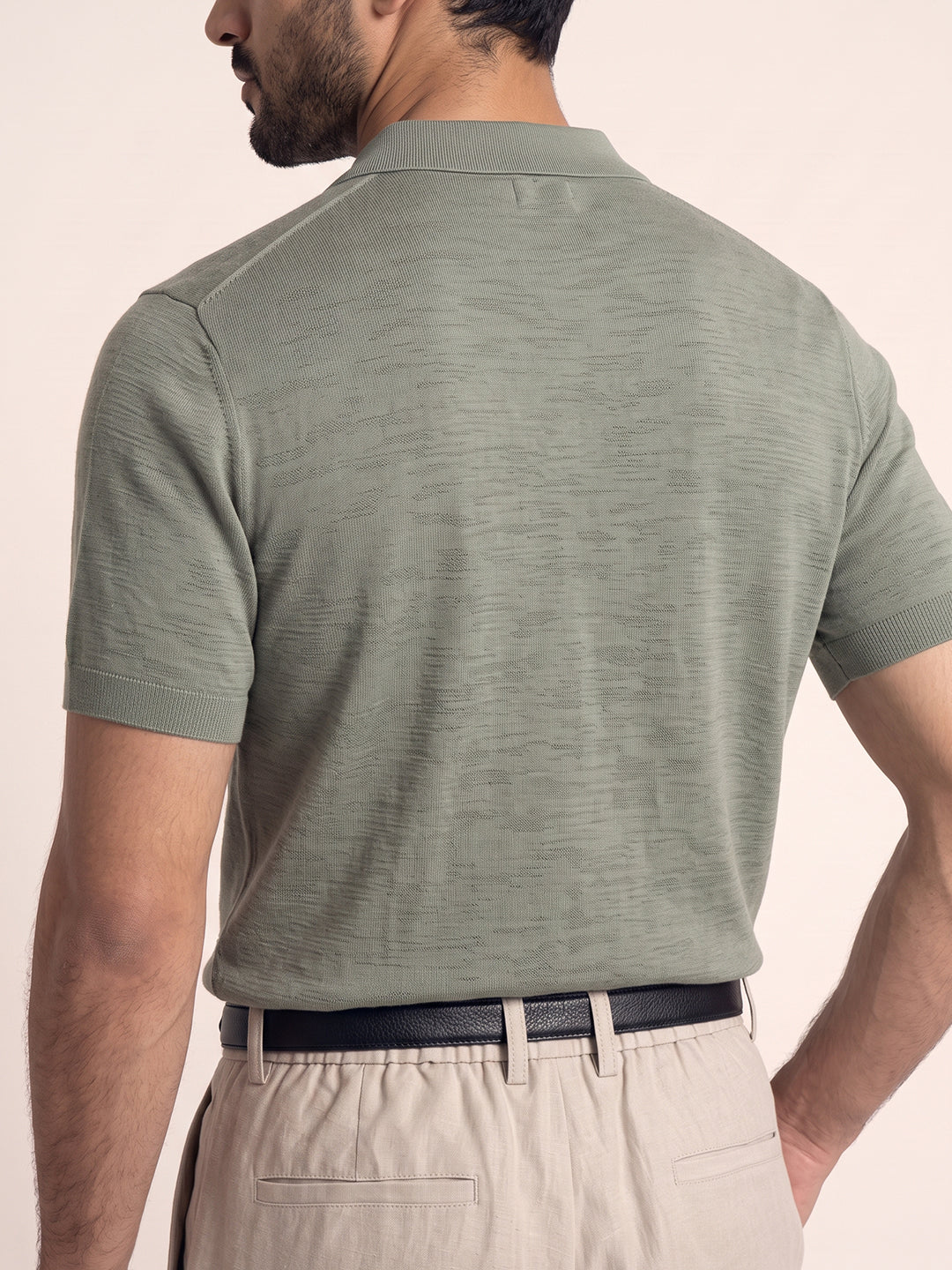 #soft-sage_group A man shown from the back wearing a short-sleeve knit polo shirt in a muted sage green color, paired with beige trousers and a black belt, against a solid light beige background.