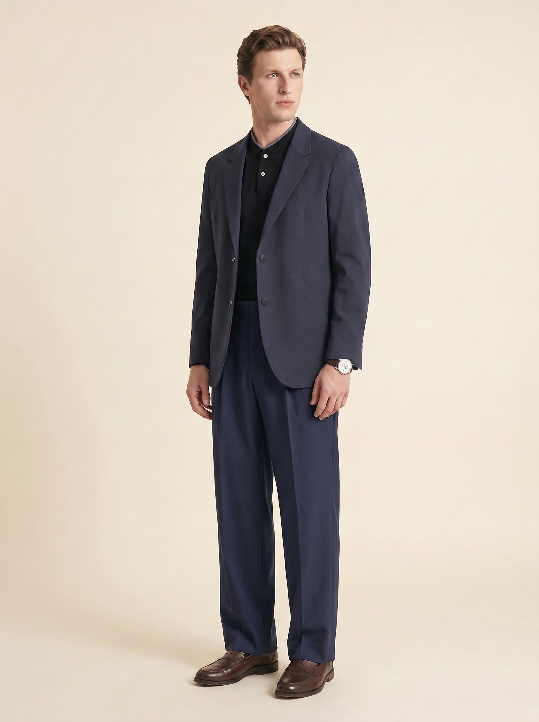 #Deep-Navy_group A man modeling a navy blue two-piece suit with a black polo shirt, brown leather loafers, and a silver watch, standing against a solid beige studio background.