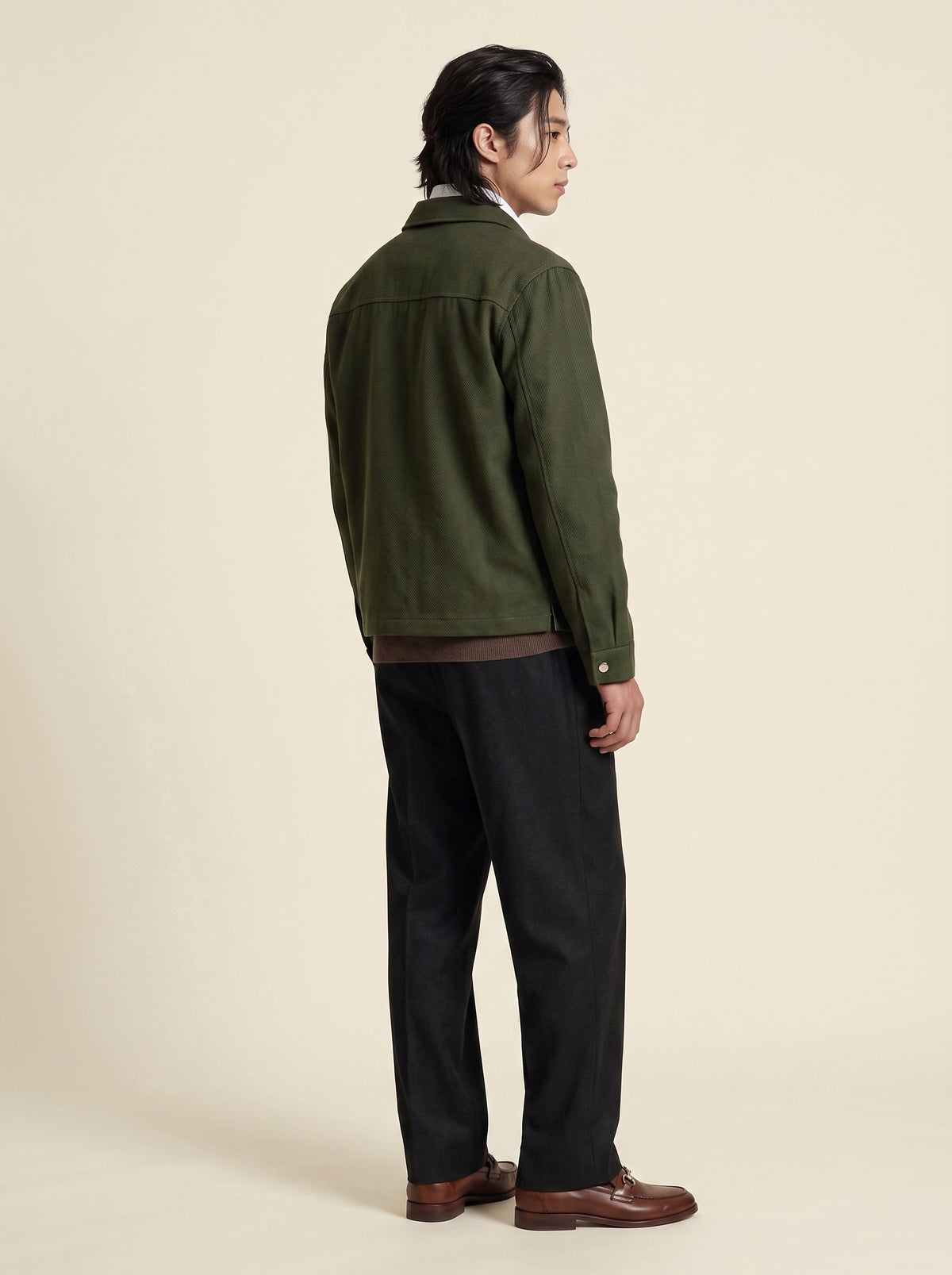 A man shown from the back wearing an olive green jacket, black trousers, and brown leather shoes, standing against a solid beige studio background.