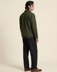 A man shown from the back wearing an olive green jacket, black trousers, and brown leather shoes, standing against a solid beige studio background.