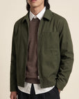 A man wearing an olive green utility jacket over a brown knit sweater, white collared shirt, and dark trousers, photographed against a plain light beige studio background.