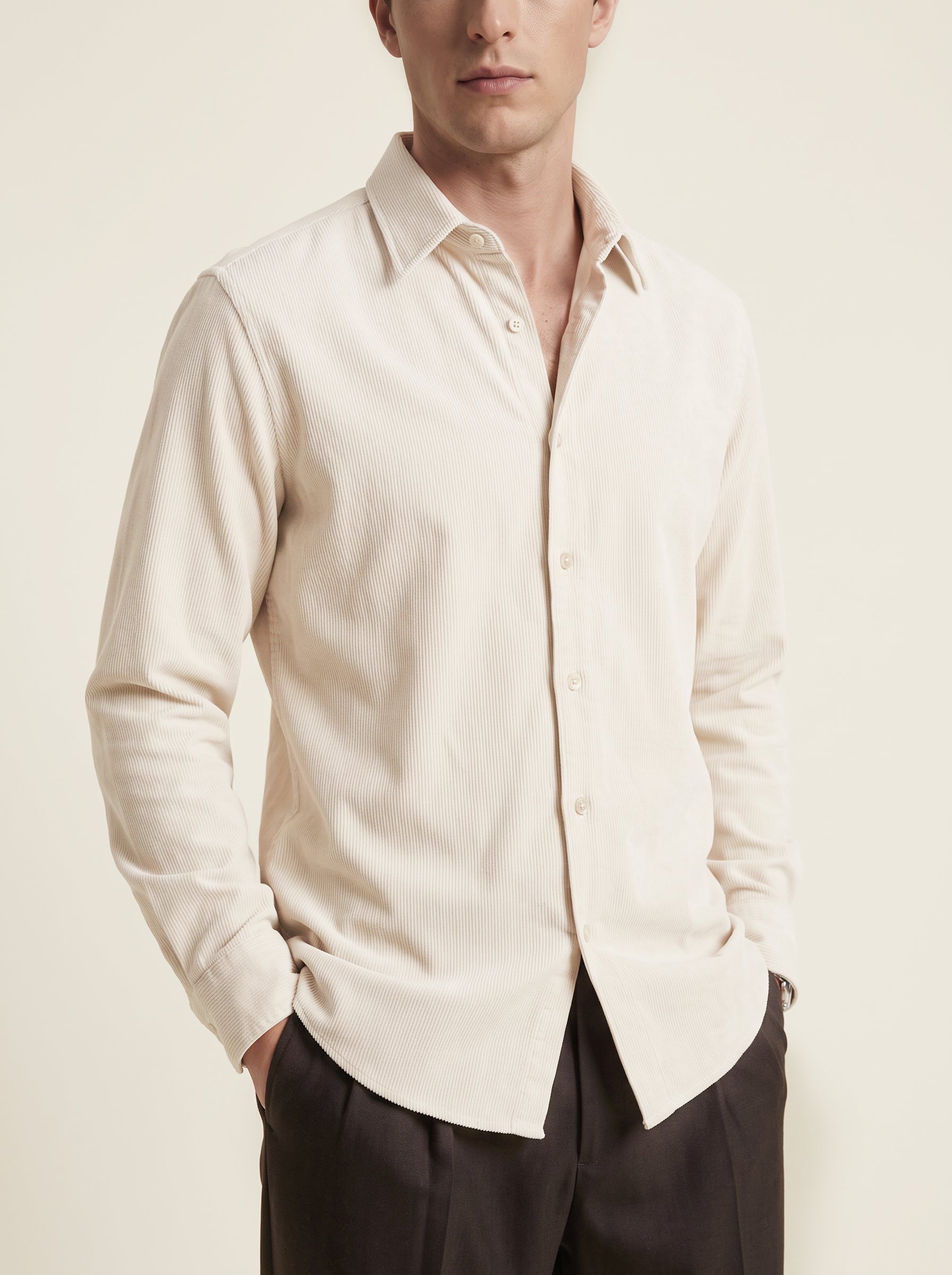 #Ecru-Ivory_group  A man wearing a light cream-colored long-sleeve linen button-down shirt and dark trousers, standing against a solid light beige background.