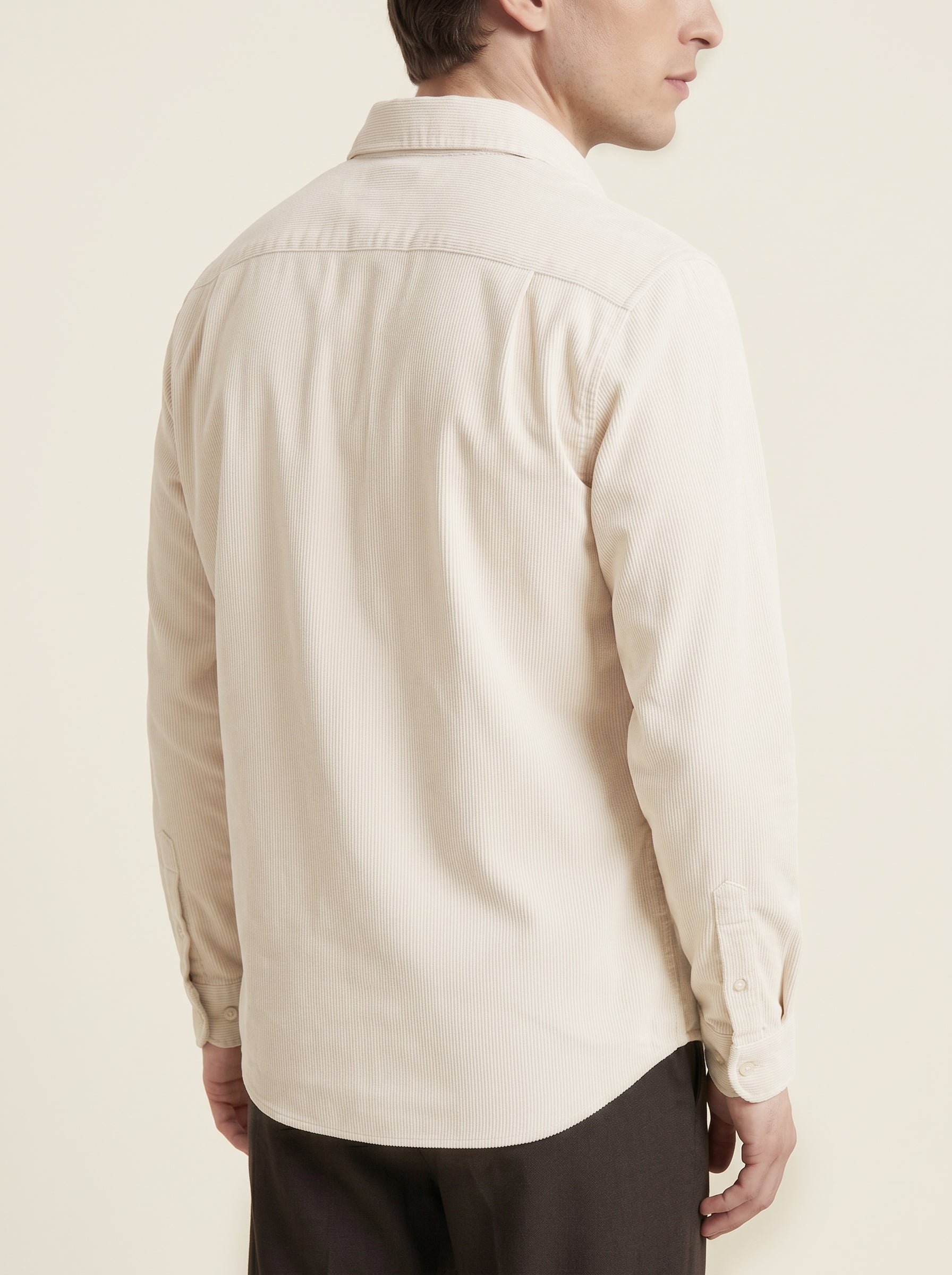 #Ecru-Ivory_group  A man shown from the back wearing a light beige long-sleeve button-up shirt and dark trousers, photographed against a plain cream background.