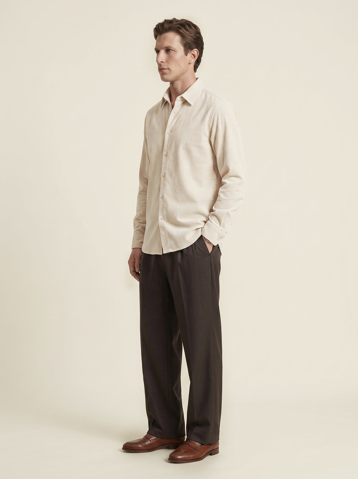 #Ecru-Ivory_group A man wearing a light beige long-sleeve button-down shirt and dark brown trousers, standing in profile against a solid cream background.