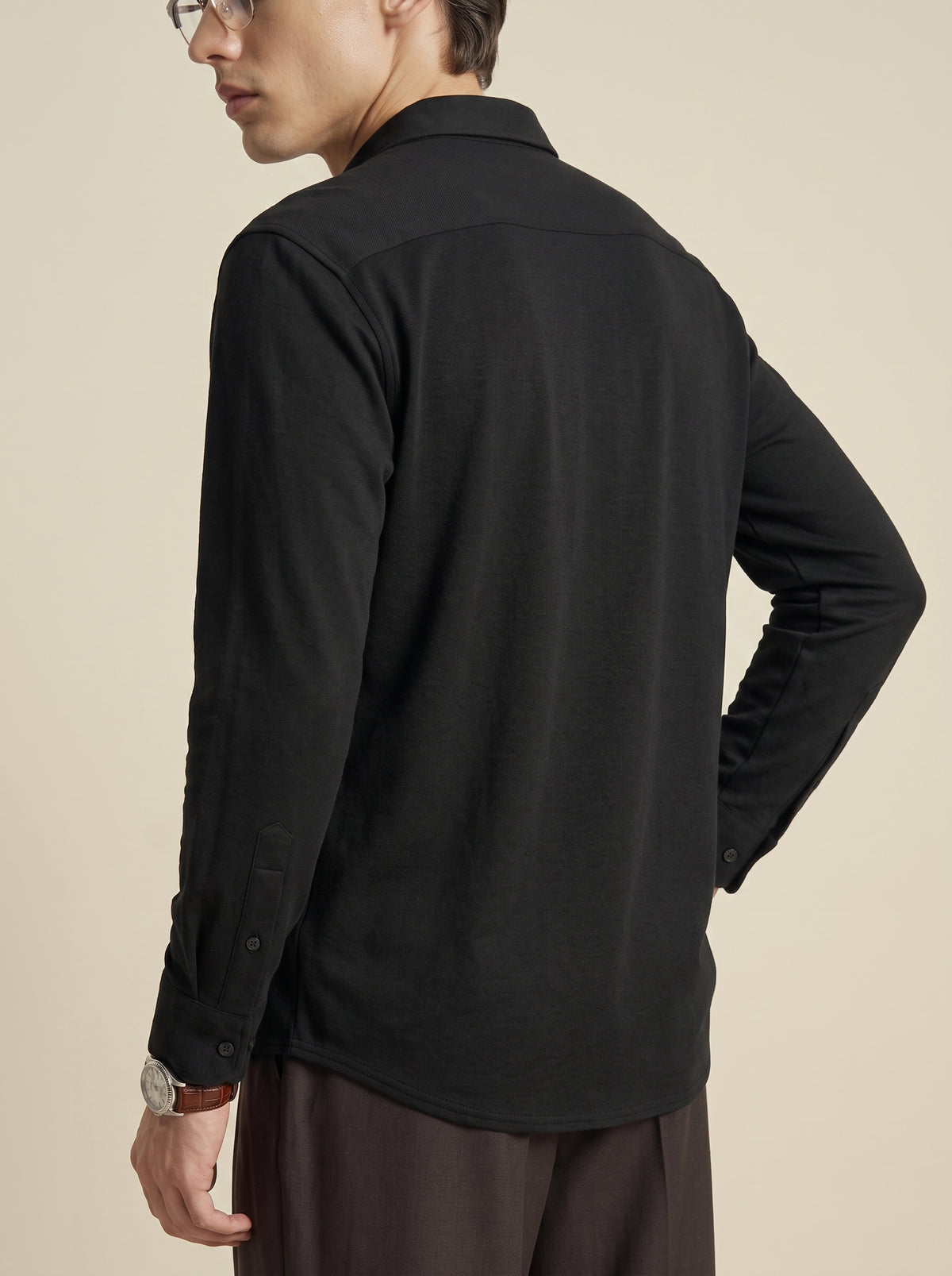 #Jet-Black_group A man wearing a black long-sleeve button-up shirt and dark brown trousers, shown from a side-back angle against a plain beige studio background.