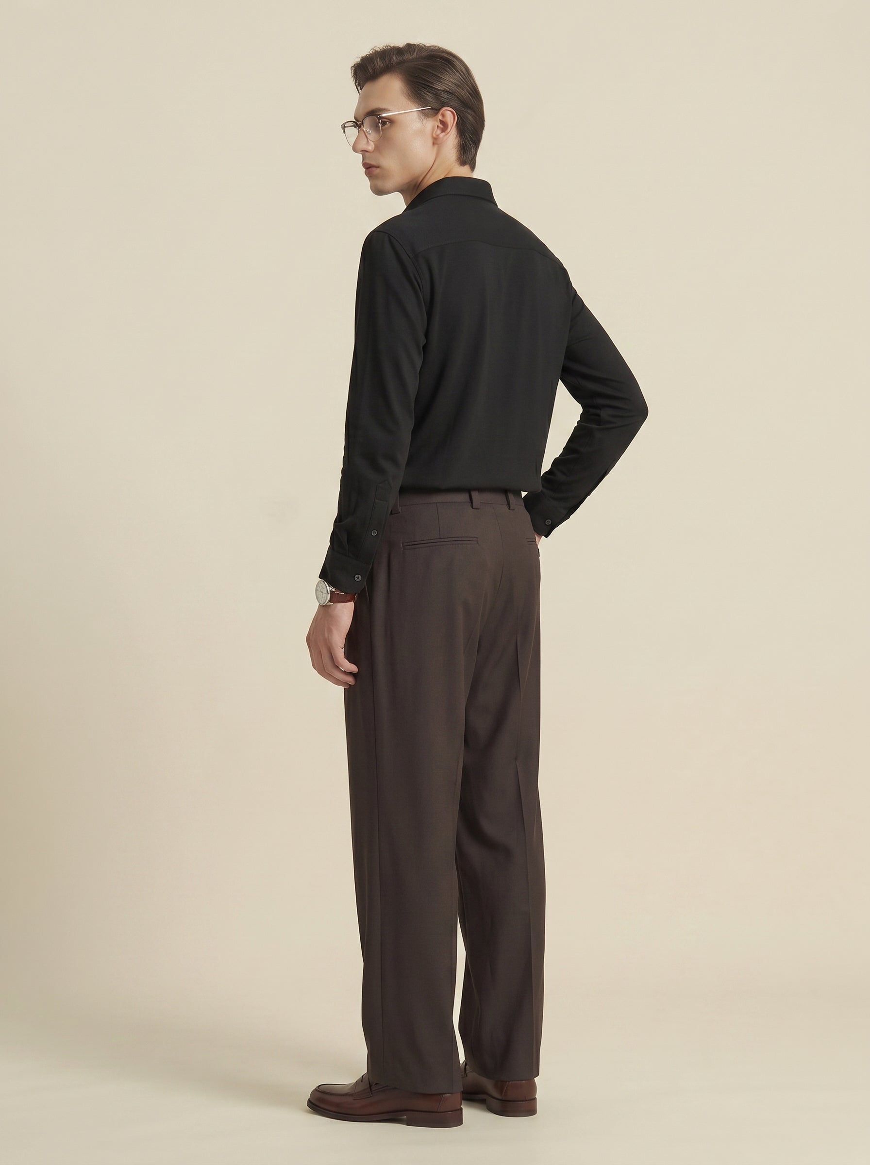 #Jet-Black_group A man wearing a black long-sleeve button-up shirt and dark brown trousers, shown from a side-back angle against a plain beige studio background.