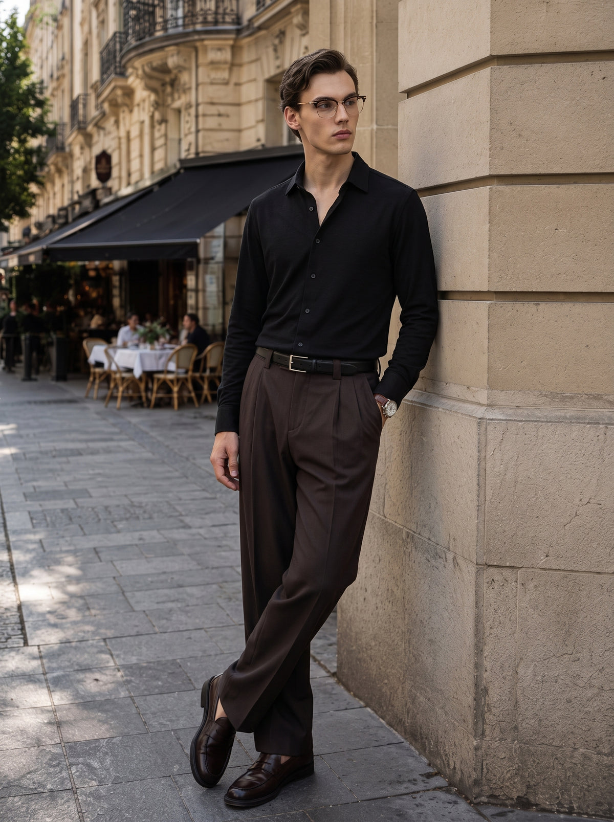 #Jet-Black_group A man wearing a black long-sleeve button-down shirt, brown trousers, and brown leather loafers leans against a beige stone building on a city sidewalk.