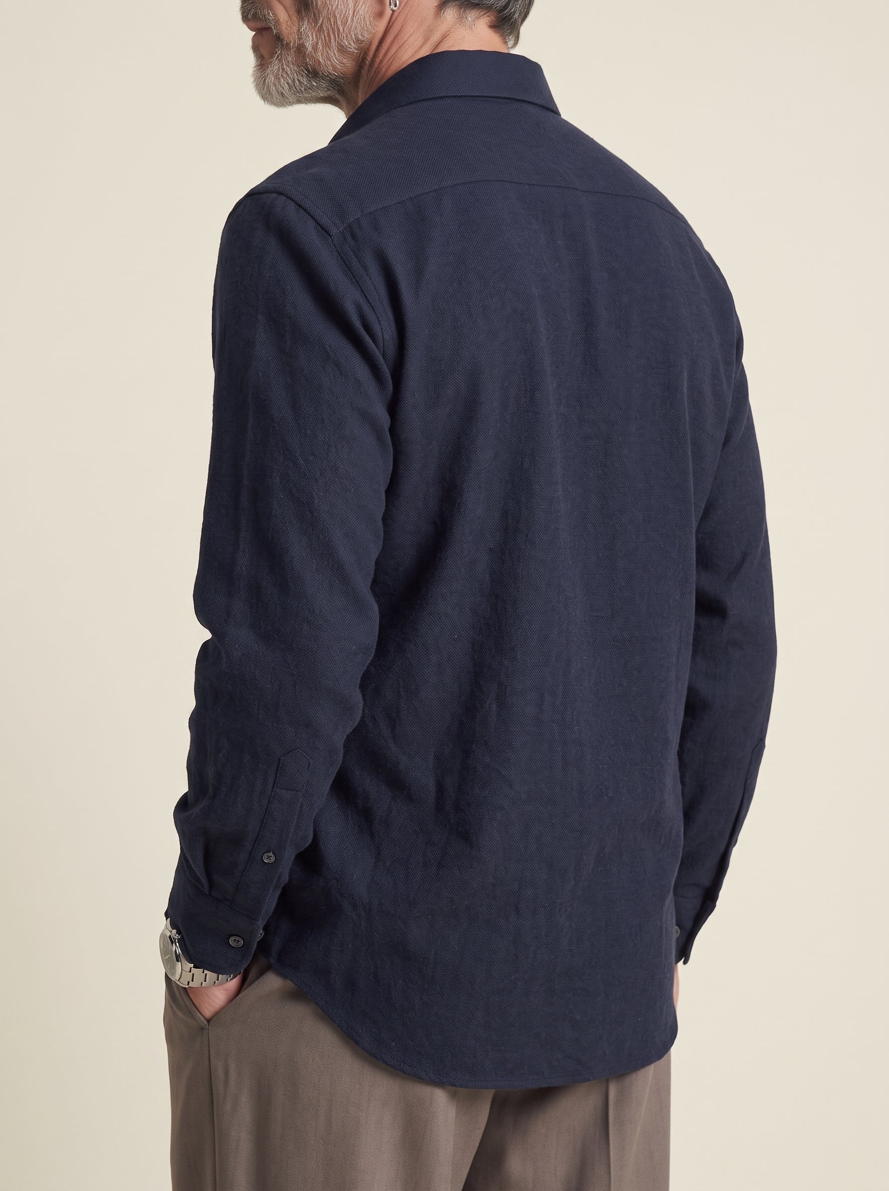 #Blue-Navy_group A man wearing a dark navy blue long-sleeve linen shirt, shown from a side-back angle against a solid light beige background.
