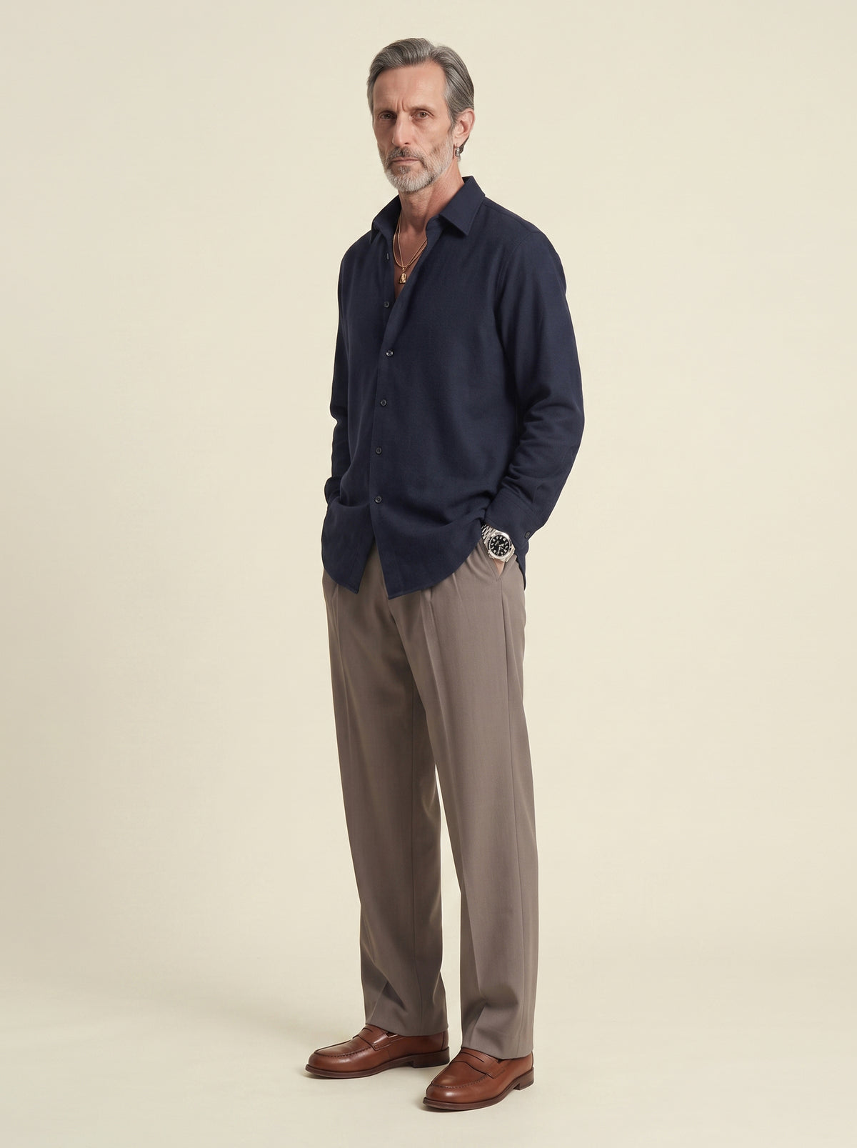 #Blue-Navy_group A man wearing a dark navy long-sleeve button-up shirt, taupe-brown dress pants, and brown leather loafers, standing against a solid light beige studio background.