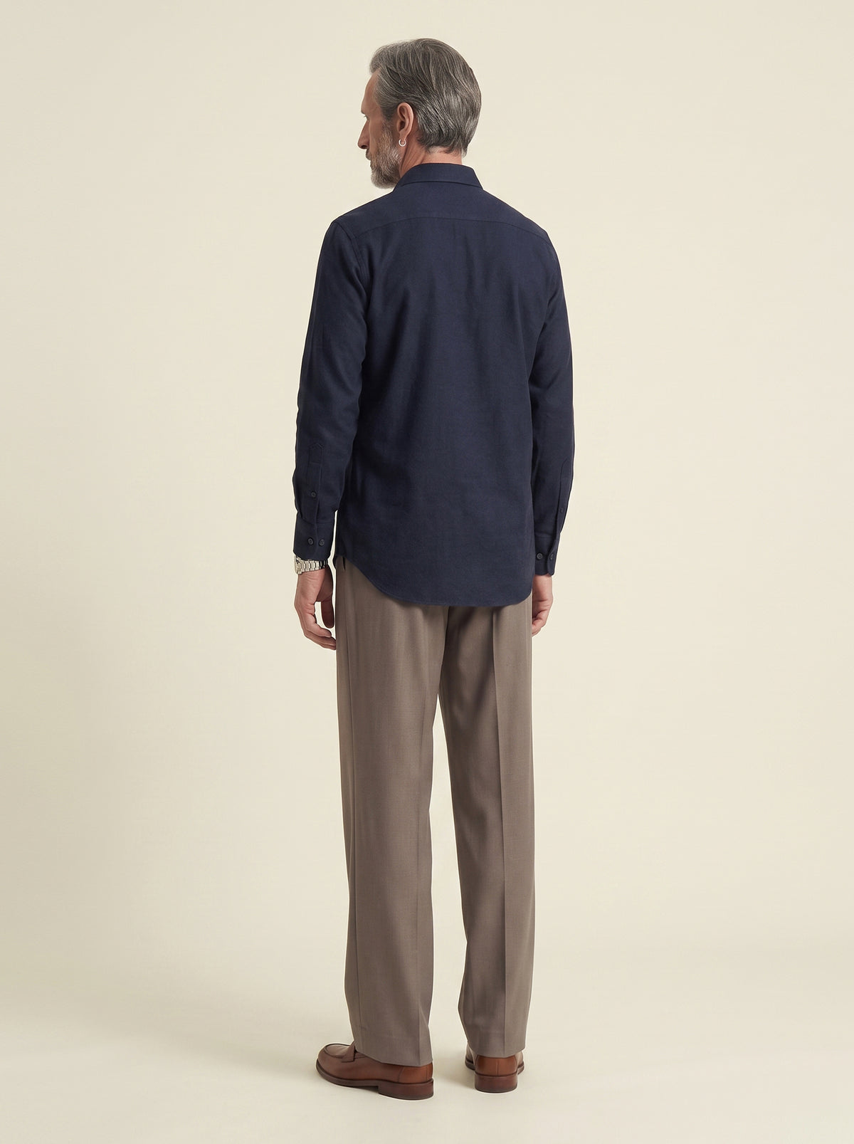 #Blue-Navy_group A man viewed from the back wearing a dark navy long-sleeve button-up shirt, taupe-brown trousers, and brown leather shoes, standing against a plain beige studio background.