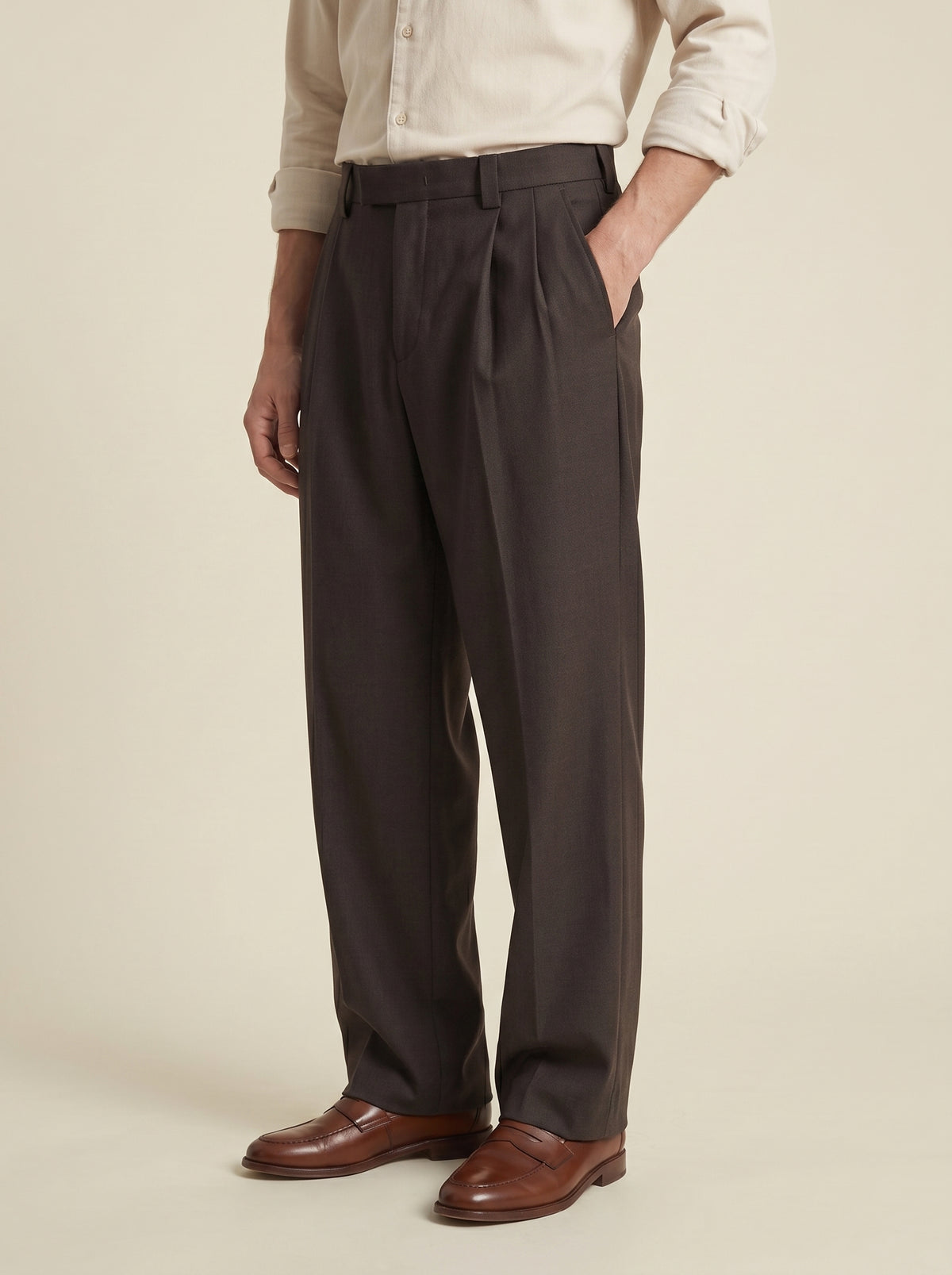 #Cedar-Brown_group A person wearing dark charcoal gray pleated trousers, a light beige shirt, and brown leather dress shoes, standing against a plain beige studio background.
