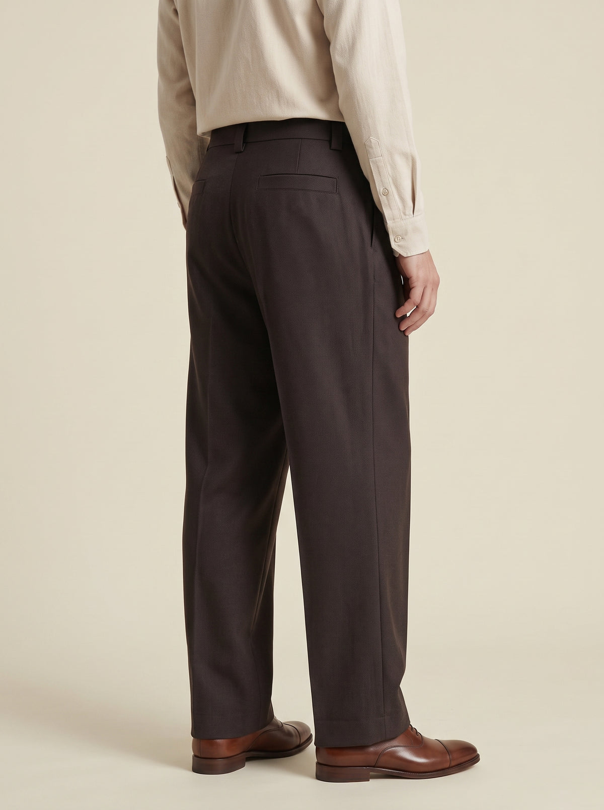 #Cedar-Brown_group A man shown from the back wearing dark charcoal gray dress pants, a light beige shirt, and brown leather shoes, set against a solid warm beige studio background.