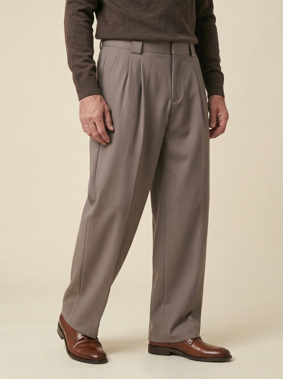 #Espresso-Brown_group A man wearing taupe pleated dress pants, a dark brown sweater, and brown leather dress shoes, standing against a solid beige studio background.