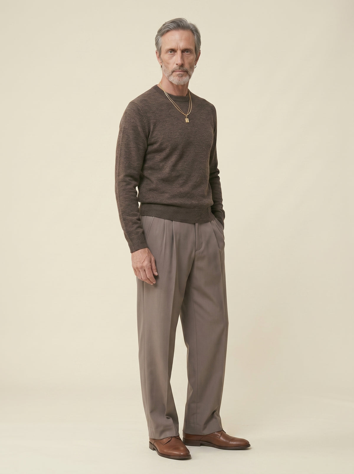 #Espresso-Brown_group A man wearing a dark brown knit crewneck sweater, taupe pleated trousers, and brown leather shoes, standing against a solid light beige background.