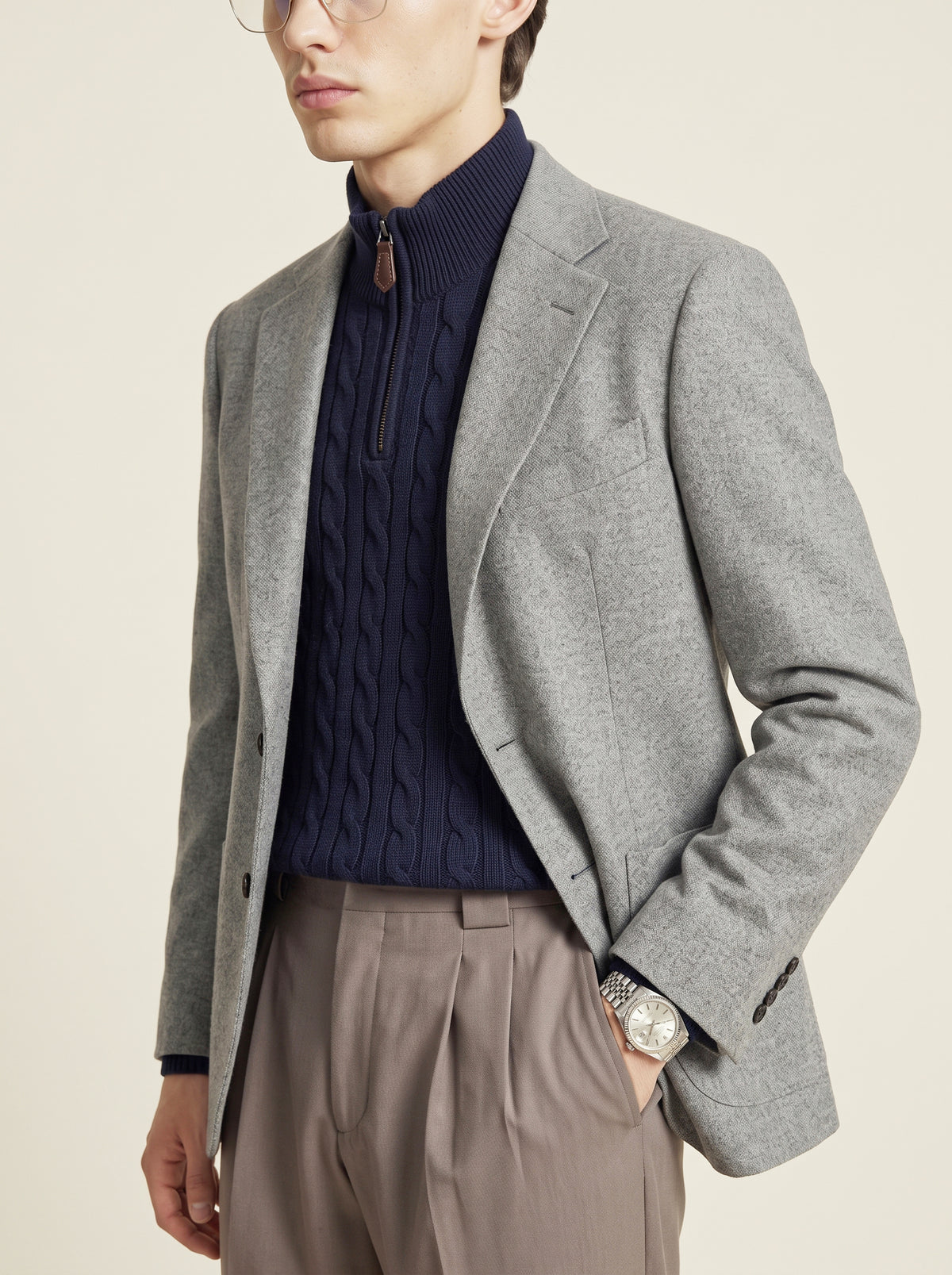 #Charcoal-Grey_group A man wearing a light gray wool blazer over a navy blue cable-knit mock-neck sweater, paired with taupe trousers, photographed against a solid light beige background.