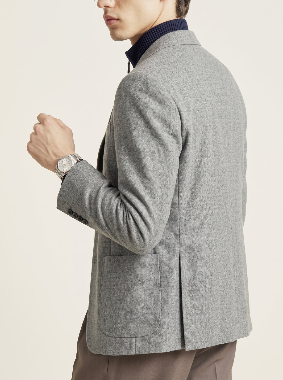 #Charcoal-Grey_group A man shown from the side wearing a light gray heathered wool blazer over a dark navy ribbed turtleneck sweater, paired with brown trousers and a silver watch, against a plain off-white background.