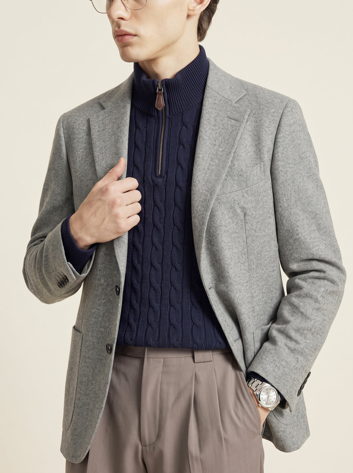 #Charcoal-Grey_group A man wearing a light gray wool blazer over a navy blue cable-knit quarter-zip sweater, paired with taupe trousers, against a solid light beige background.