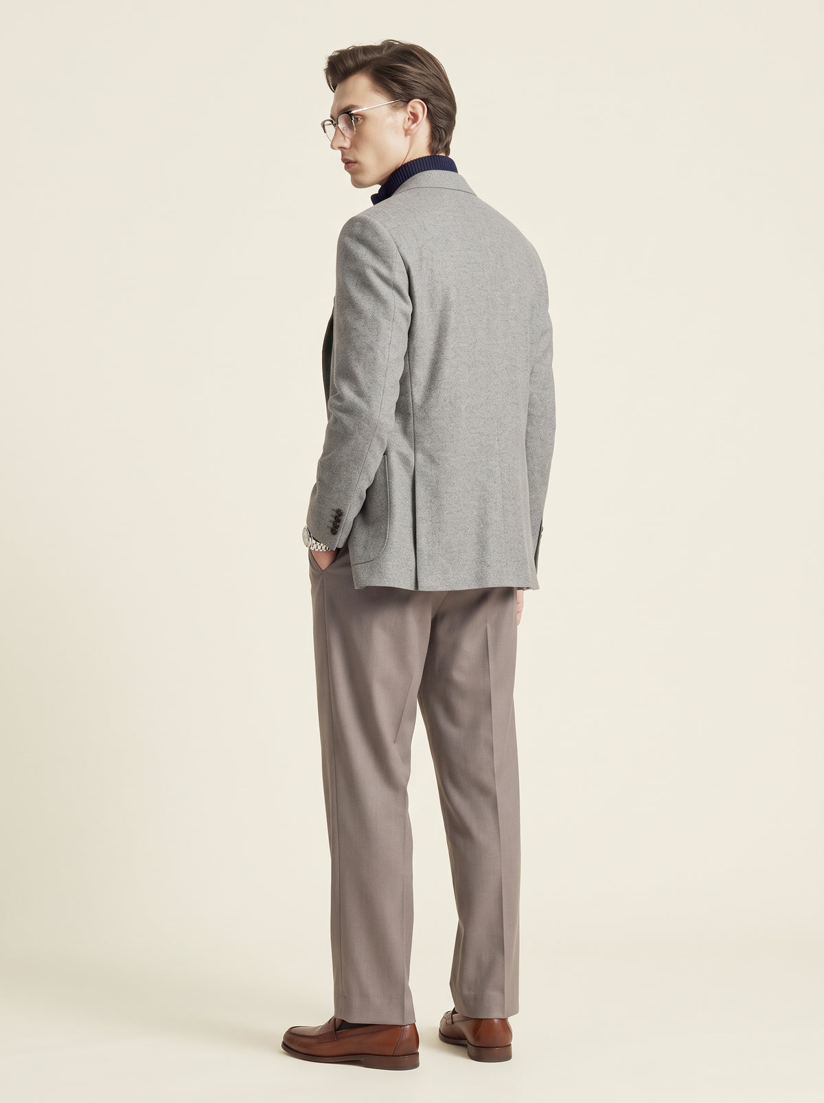 #Charcoal-Grey_group A man wearing a light gray blazer, taupe trousers, and brown leather loafers, photographed from the back against a plain cream studio background.