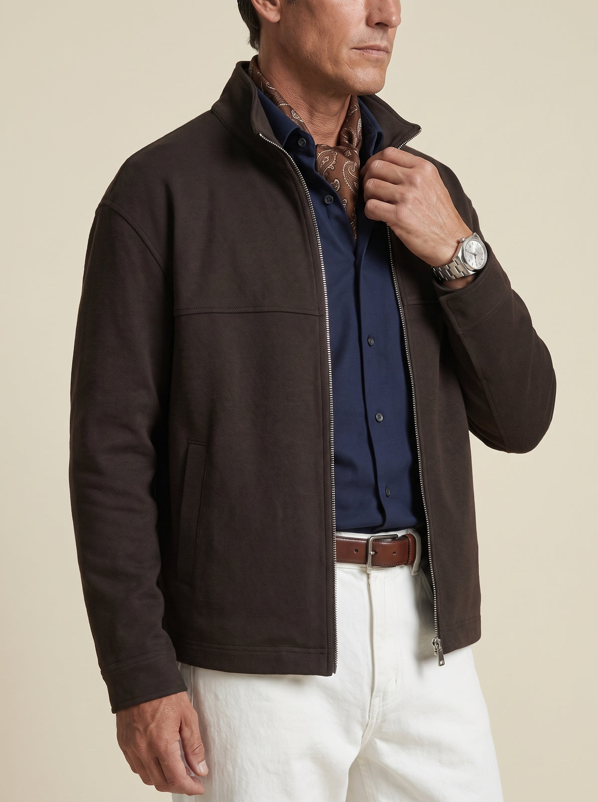 #Cedar-Brown_group A man wearing a dark brown zip-up fleece jacket over a navy blue button-down shirt, white trousers, and a brown belt, photographed against a solid beige background.