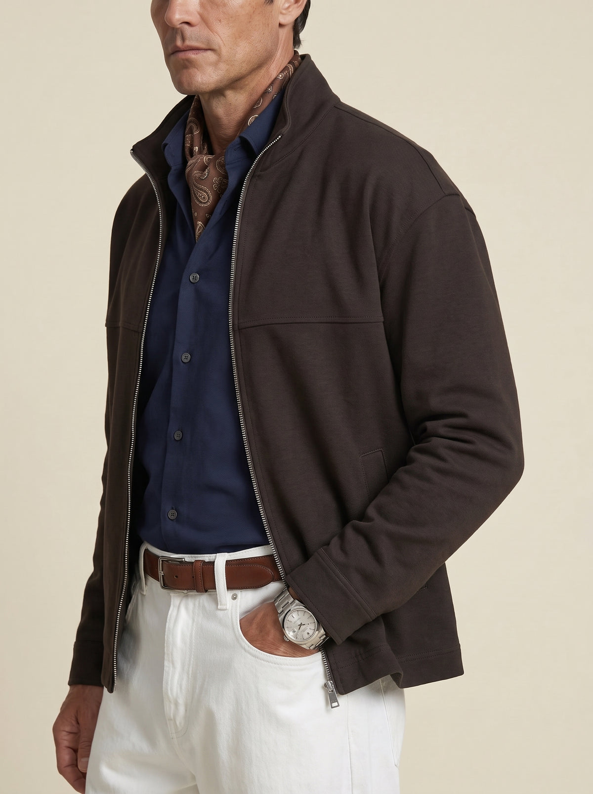 #Cedar-Brown_group A man wearing a dark brown zip-up jacket over a navy blue button-down shirt, white trousers, and a brown belt, photographed against a plain beige studio background.