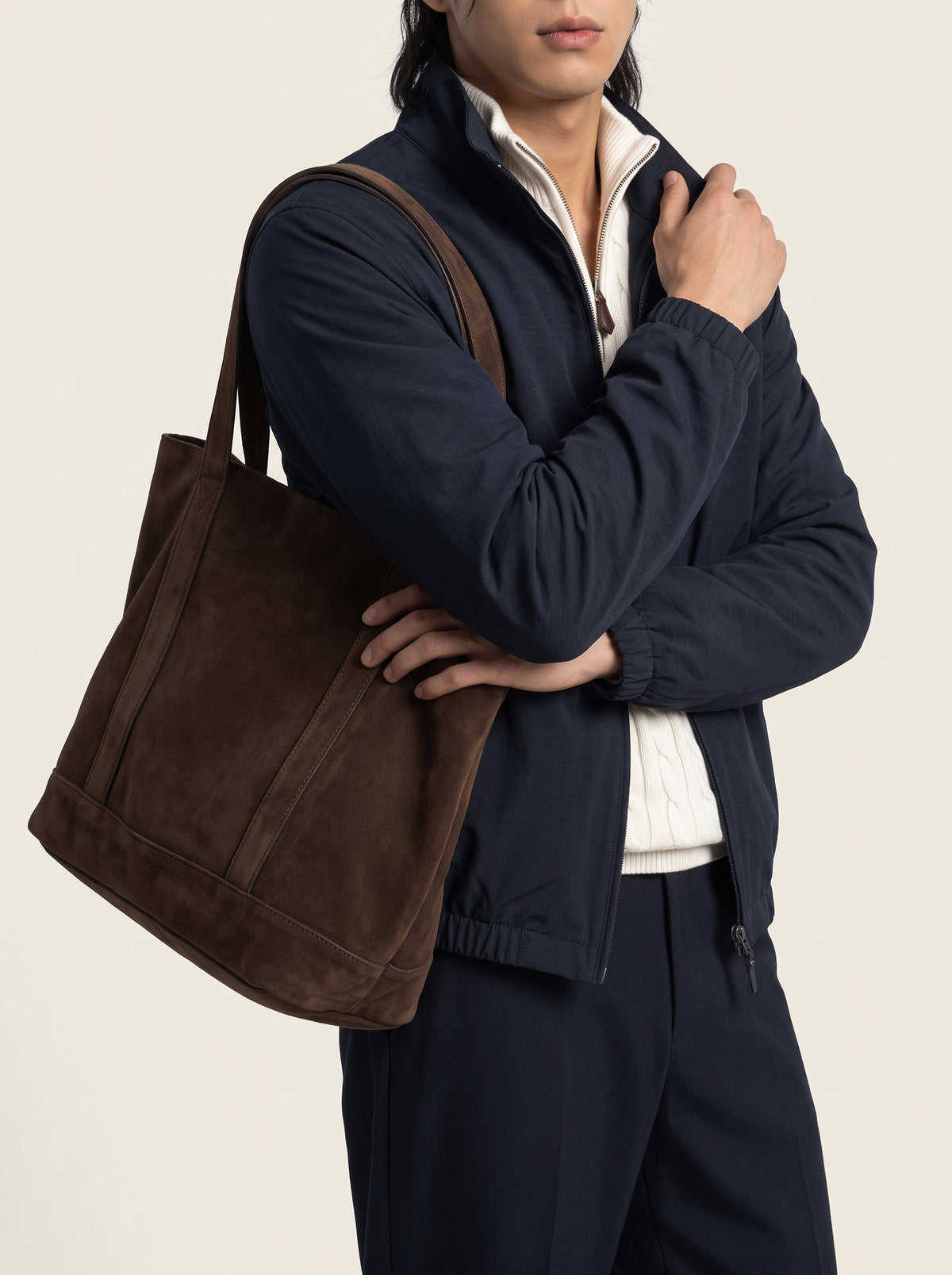 #Blue-Navy_group A man wearing a navy jacket and matching pants carries a large brown suede tote bag over his shoulder, photographed against a plain light beige studio background.