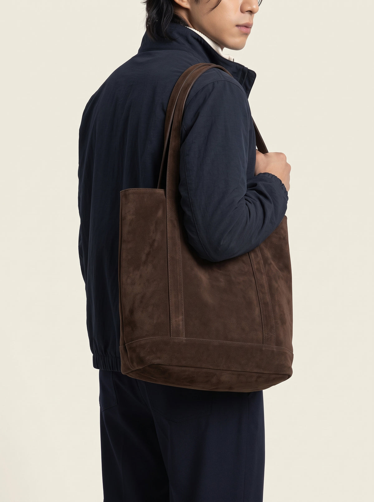 #Blue-Navy_group A person wearing a dark navy jacket and pants carries a large brown suede tote bag over the shoulder, shown from a side-back angle against a plain light beige studio background.