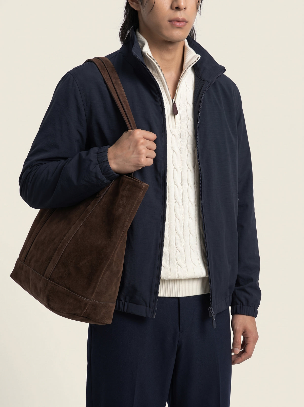 #Blue-Navy_group A man wearing a navy blue lightweight jacket over a cream cable-knit zip-up sweater, carrying a large brown suede tote bag on his shoulder, against a plain light beige background.