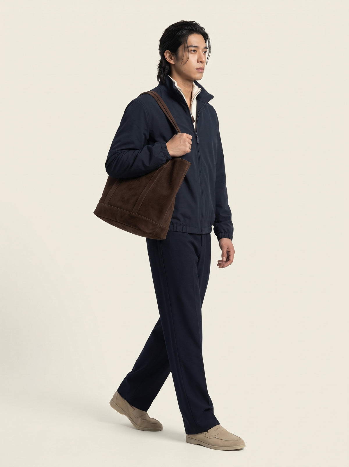 #Blue-Navy_group A man wearing a navy blue zip-up jacket and matching navy pants, carrying a large brown suede shoulder bag, walking against a light beige studio background.
