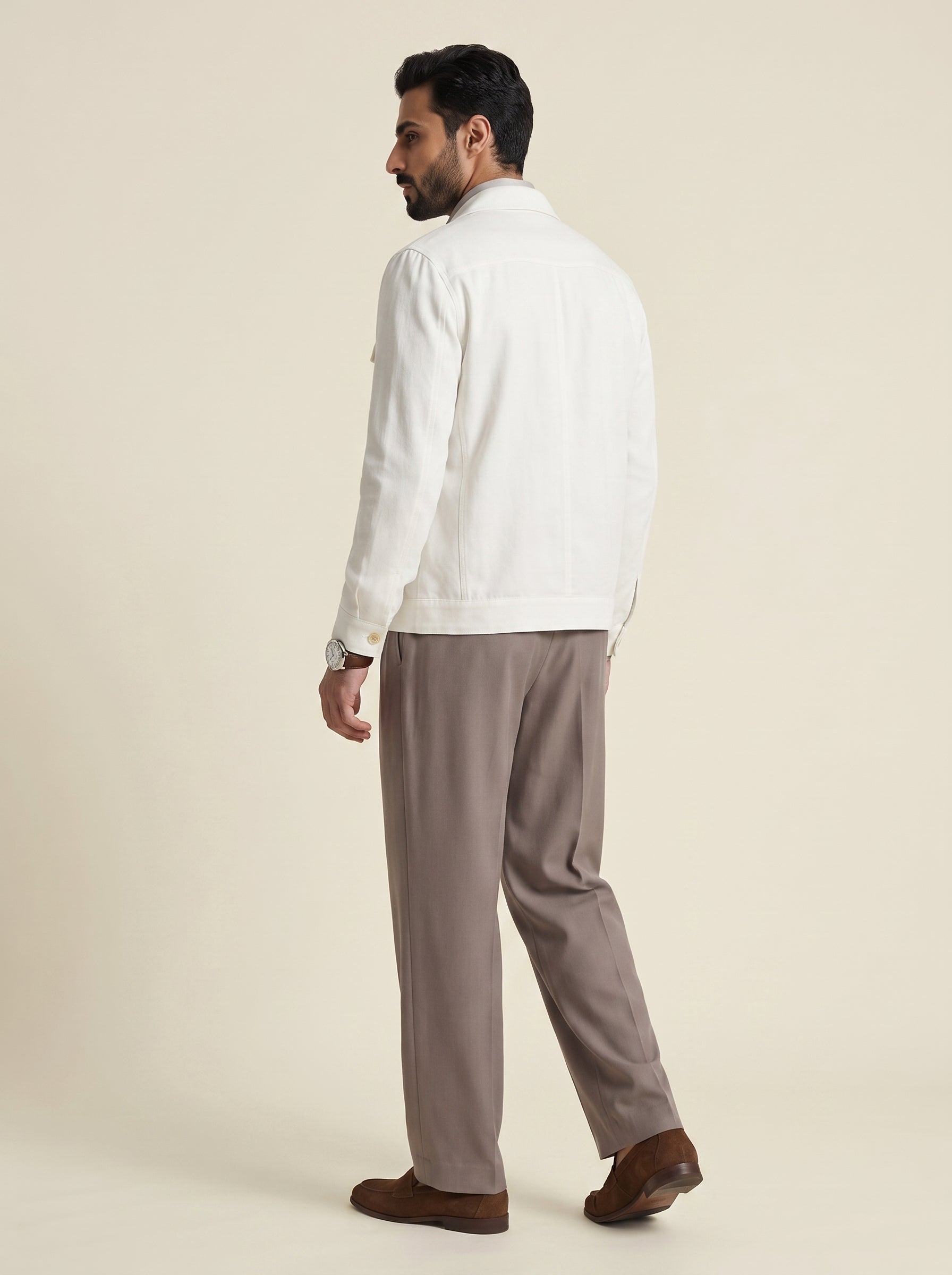 #Pure-White_group A man shown from the back wearing a white jacket, taupe-brown trousers, and brown loafers, standing against a solid light beige studio background.