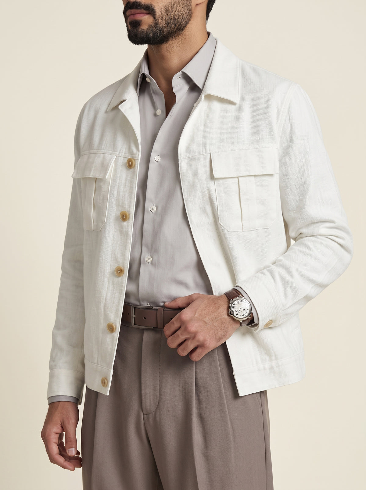 #Pure-White_group A man wearing a light cream linen jacket over a light gray button-down shirt, paired with taupe trousers and a brown belt, photographed against a solid beige background.
