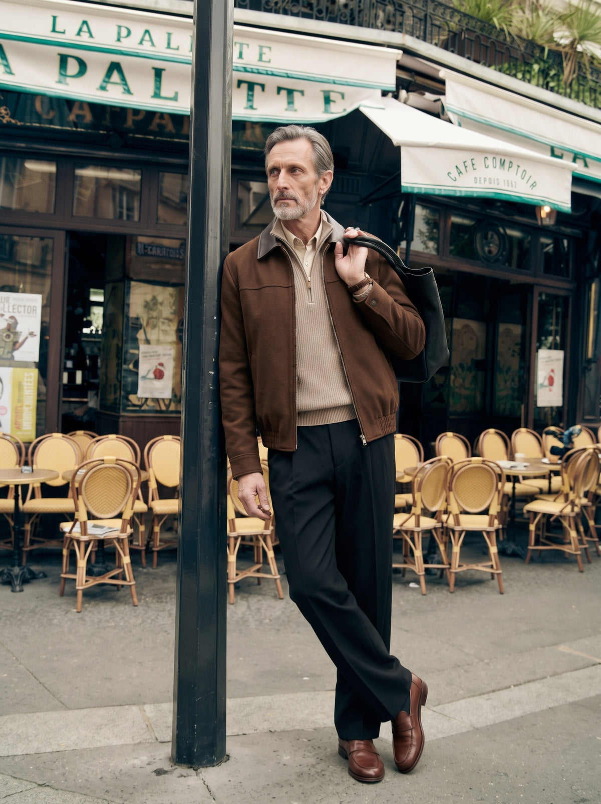#Cedar-Brown_group A man wearing a brown leather jacket, beige sweater, black trousers, and brown leather shoes stands on a Parisian street sidewalk next to a cafe, holding a black shoulder bag.