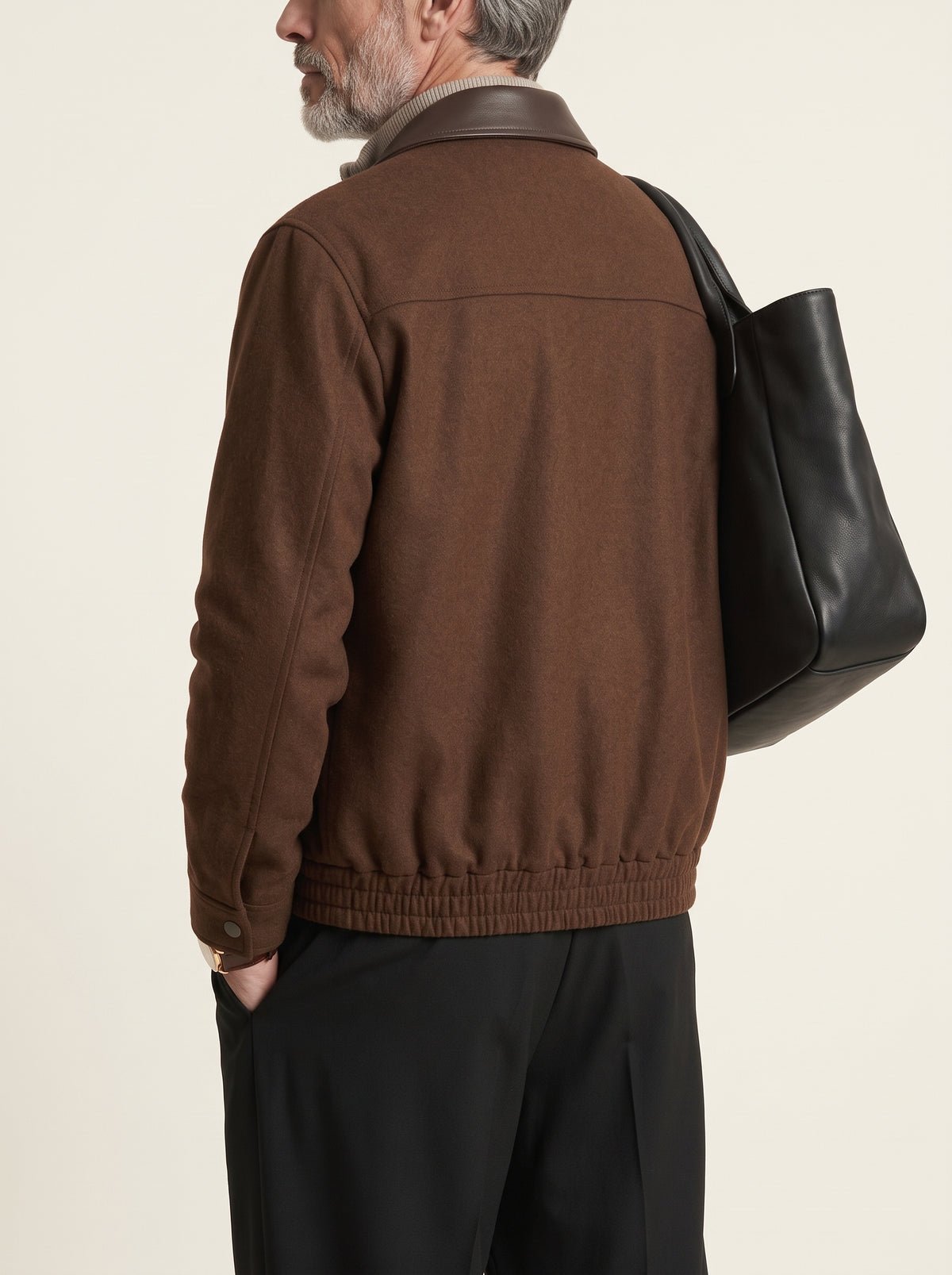 #Cedar-Brown_group A man shown from the back wearing a brown wool-blend jacket with dark brown leather trim on the collar and cuffs, paired with black trousers and carrying a large black leather shoulder bag against a light beige background.