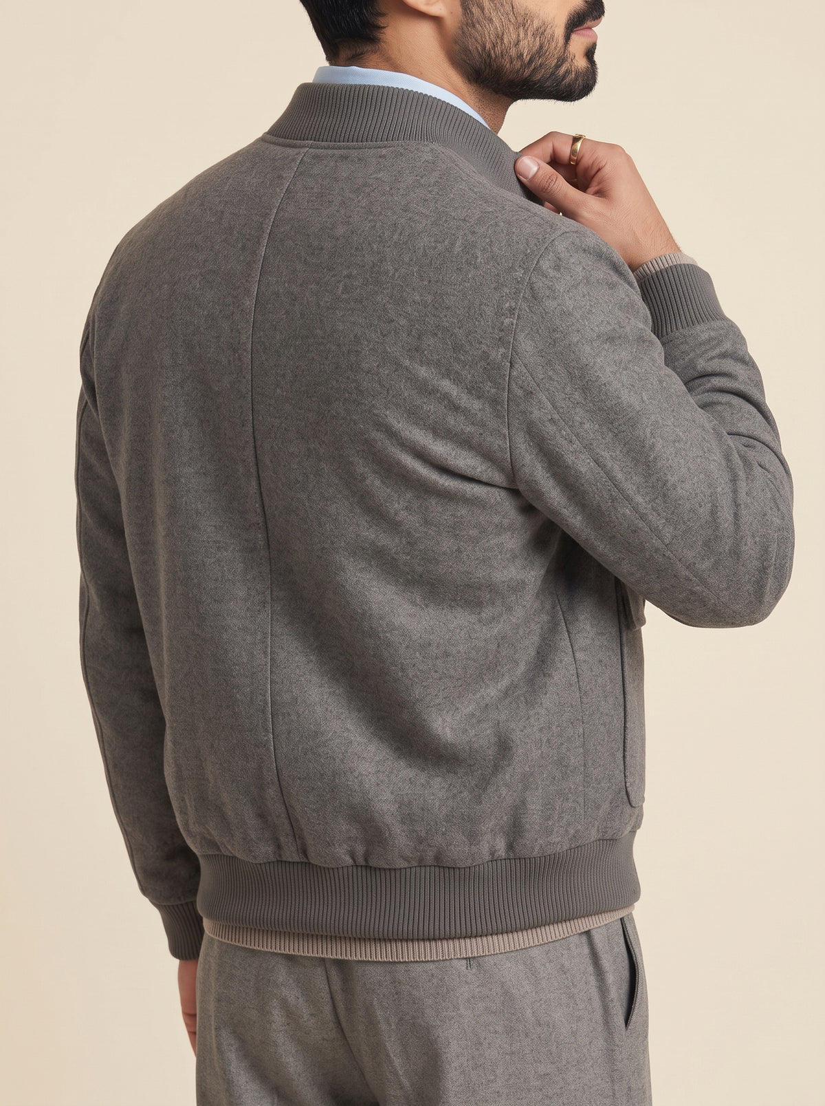 #Charcoal-Grey_group A man shown from the back wearing a heather gray knit bomber-style cardigan with ribbed collar, cuffs, and hem, layered over a light blue collared shirt. The background is a solid, warm off-white studio backdrop.