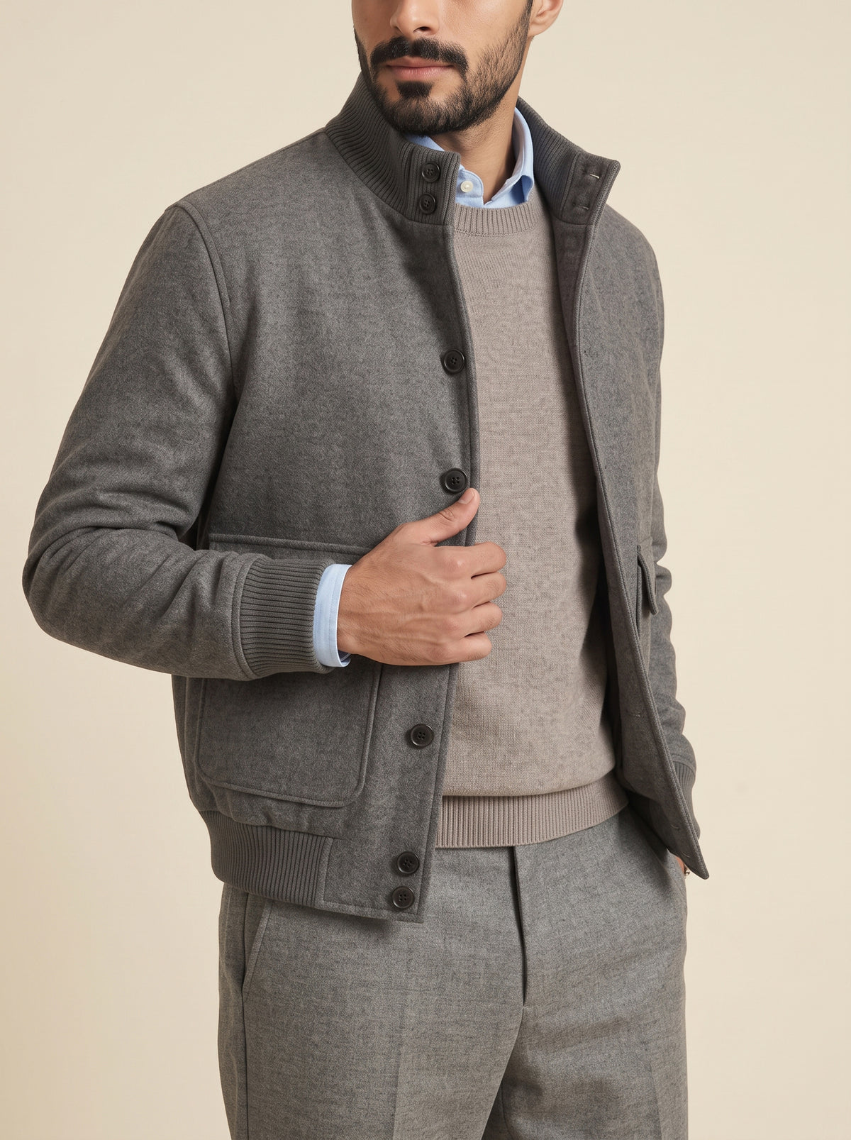 #Charcoal-Grey_group A man wearing a gray knit cardigan jacket over a taupe sweater and trousers, styled with a light blue dress shirt. The photo is cropped from the chin down against a solid beige background.