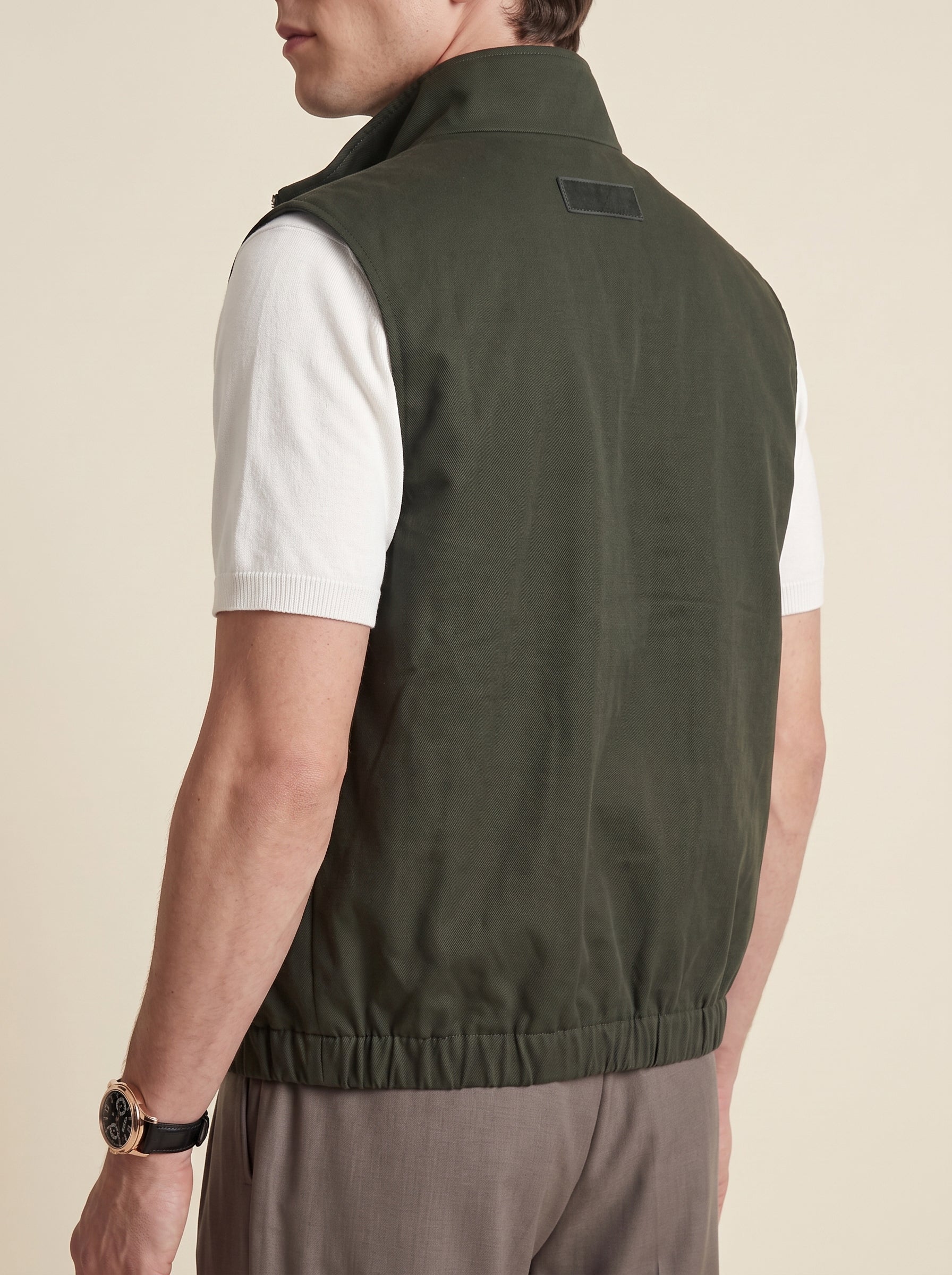#Olive-Leaf_group A man shown from the back wearing an olive green sleeveless zip vest over a white short-sleeve shirt and taupe pants, against a warm beige studio background.