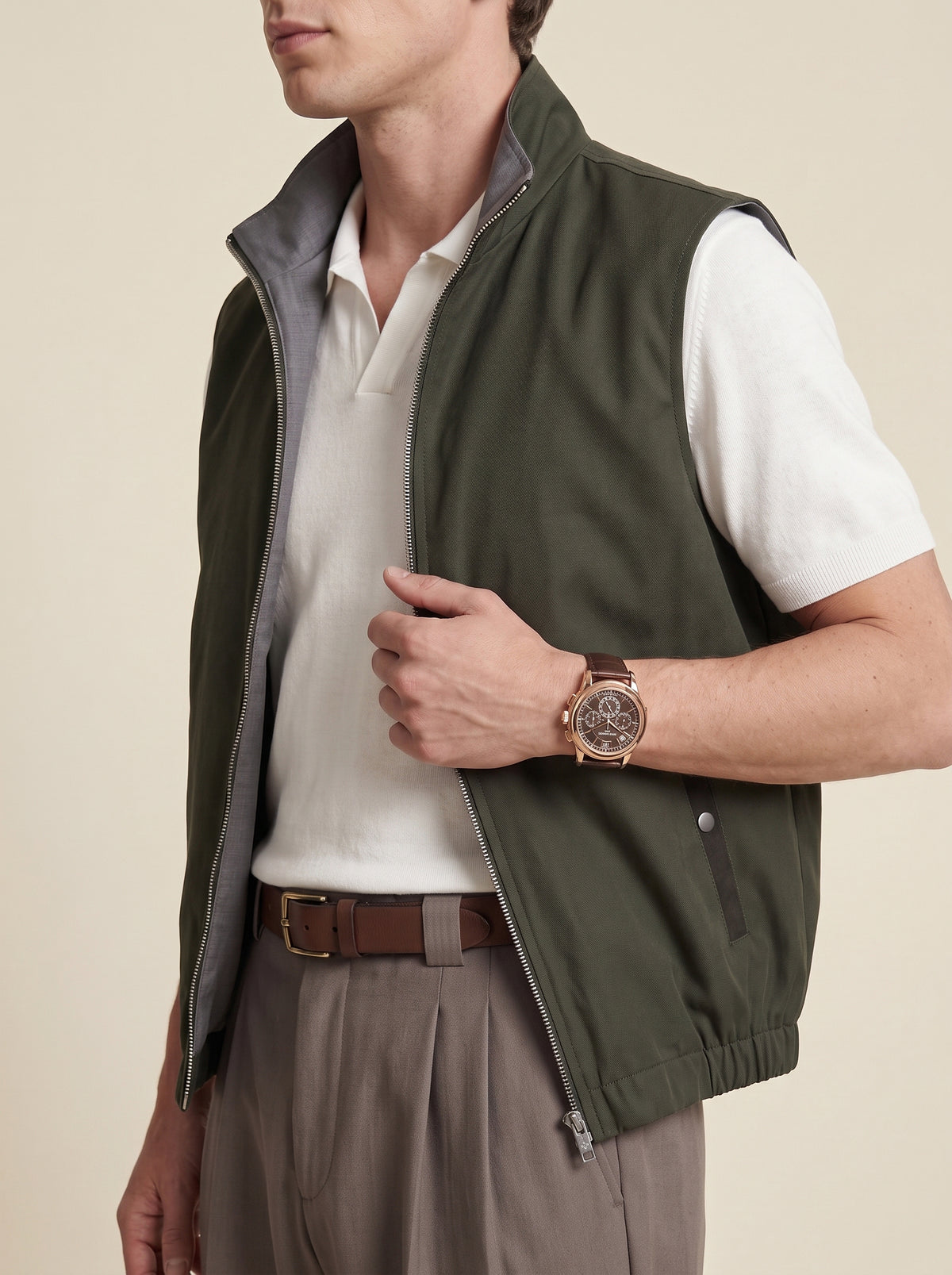 #Olive-Leaf_group A man wearing an olive green zip-up sleeveless vest over a white polo shirt and brown trousers, shown from the neck down against a plain beige studio background.