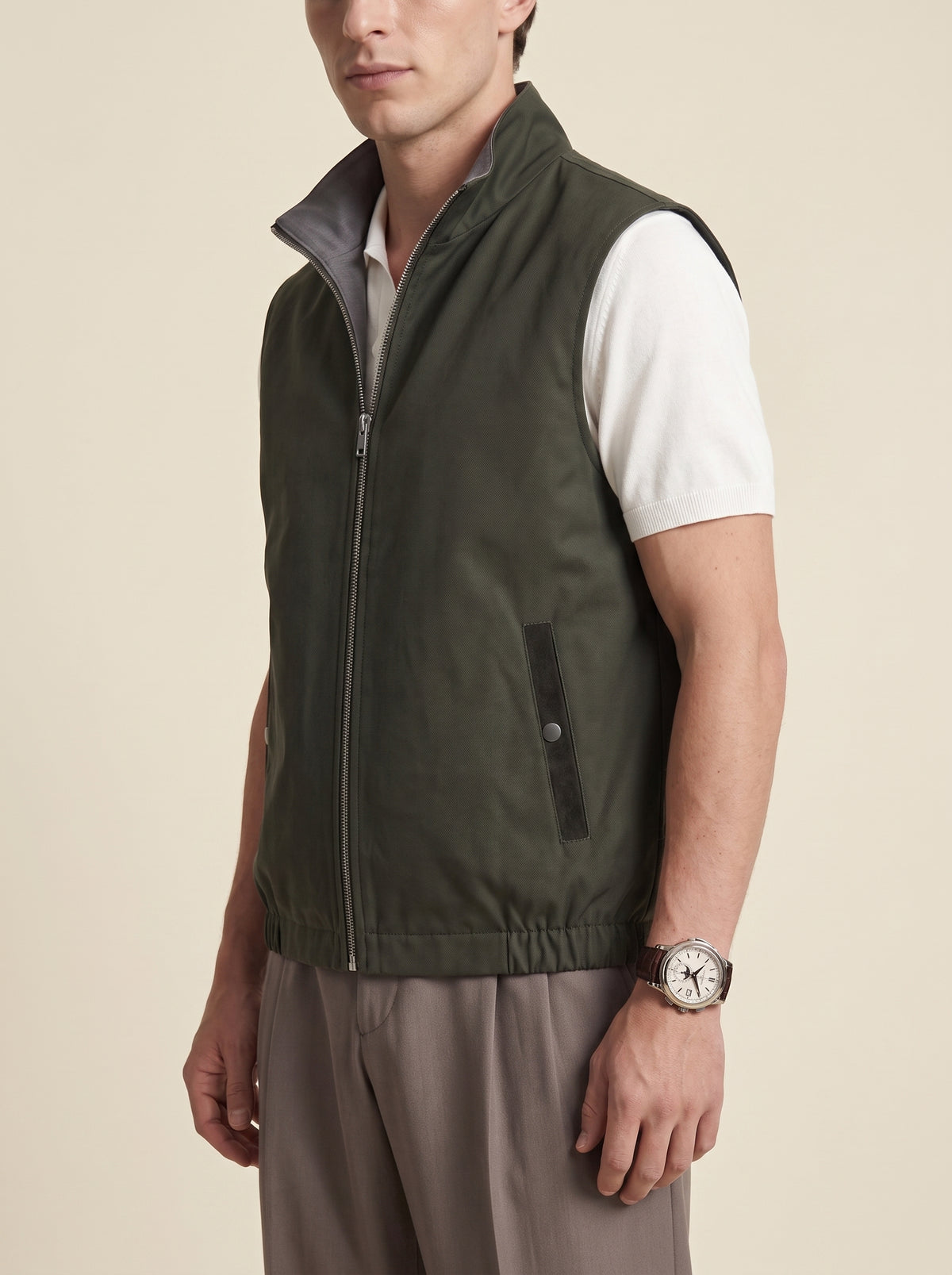 #Olive-Leaf_group A man wearing an olive green sleeveless zip-front vest over a white short-sleeve shirt and taupe pants, photographed against a plain warm beige studio background.