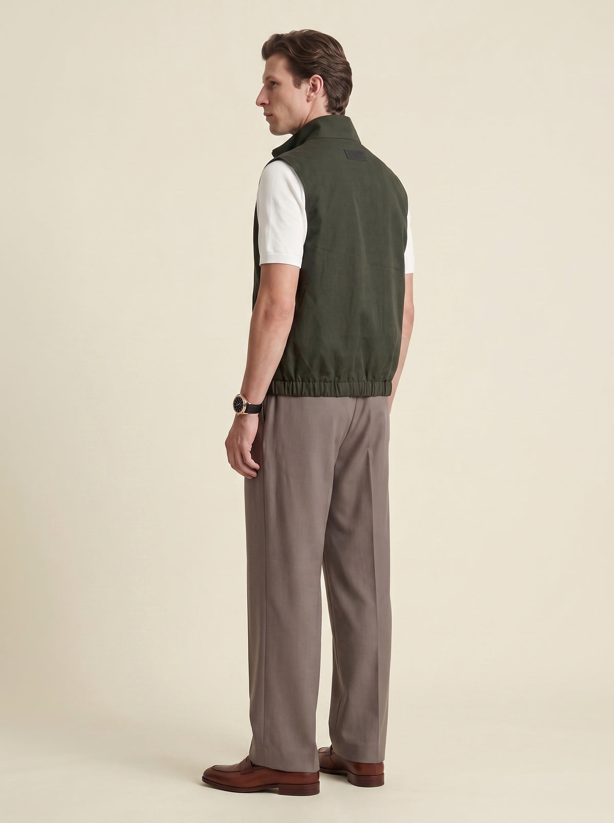 #Olive-Leaf_group A man shown from the back wearing an olive green sleeveless vest over a white short-sleeve shirt, paired with taupe trousers and brown shoes, against a solid beige background.