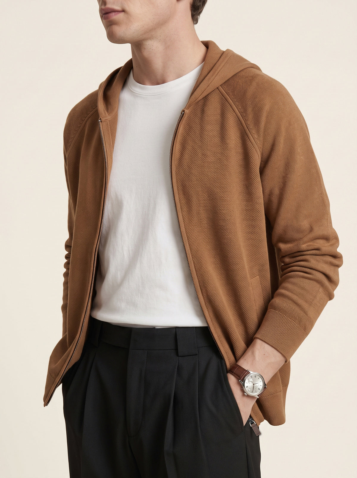 #Cedar-Brown_group A man wearing a camel brown hooded zip-up cardigan over a white t-shirt and black trousers, shown from the chin down against a light beige background.