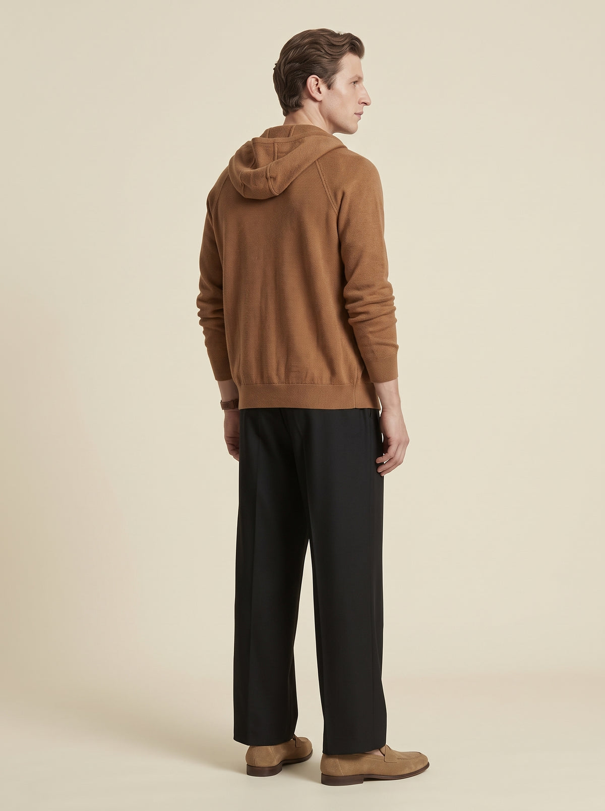 #Cedar-Brown_group A man shown from the back wearing a camel brown hooded sweater, black trousers, and tan shoes, standing against a solid light beige background.