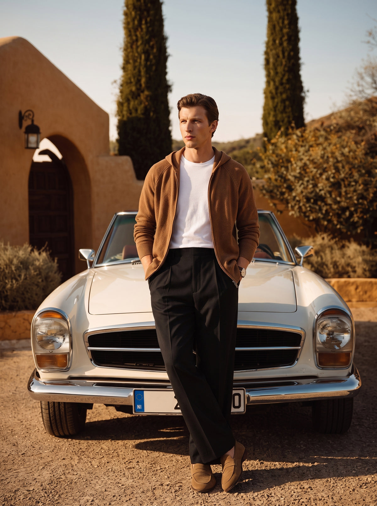#Cedar-Brown_group A man stands in front of a vintage white convertible car outdoors, wearing a brown zip-up hoodie over a white T-shirt, black trousers, and brown shoes. The setting includes a tan building, tall cypress trees, and dry hills under warm sunlight.
