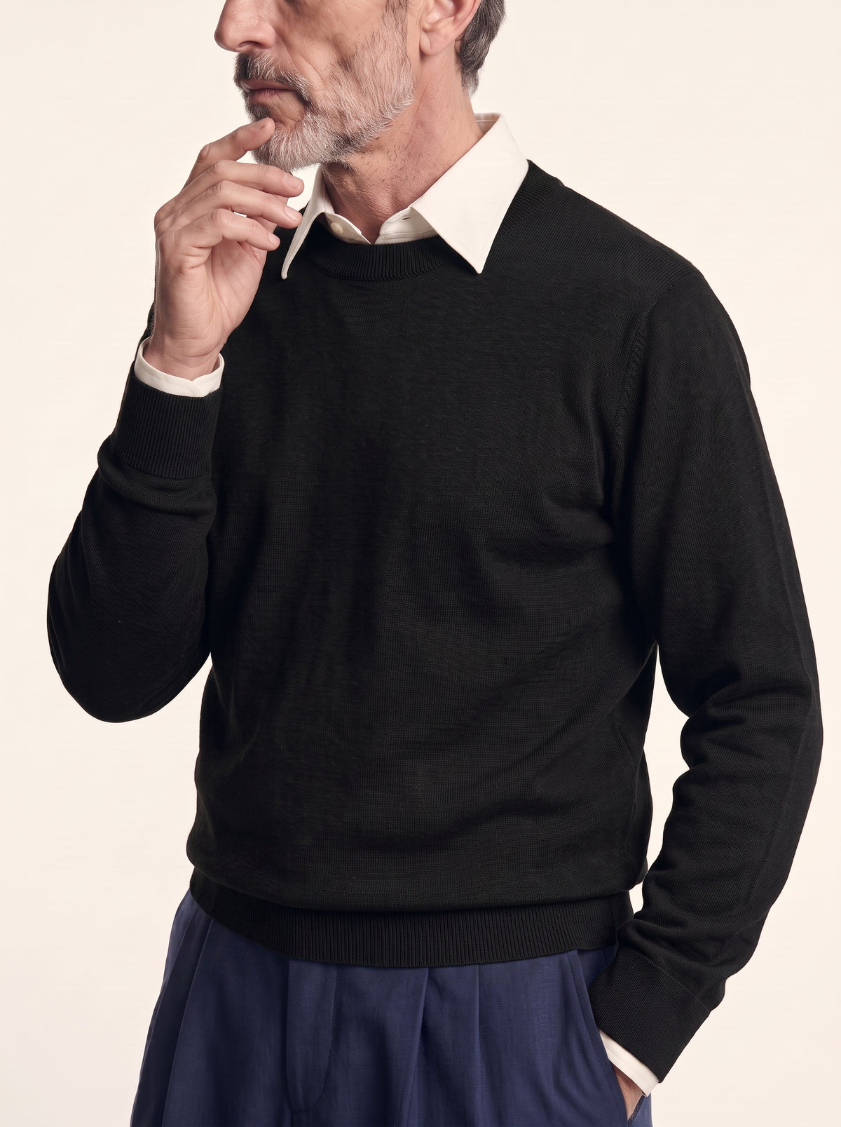 #Jet-Black_group A man wearing a black crewneck sweater over a cream collared shirt and navy trousers, posing against a light beige background.