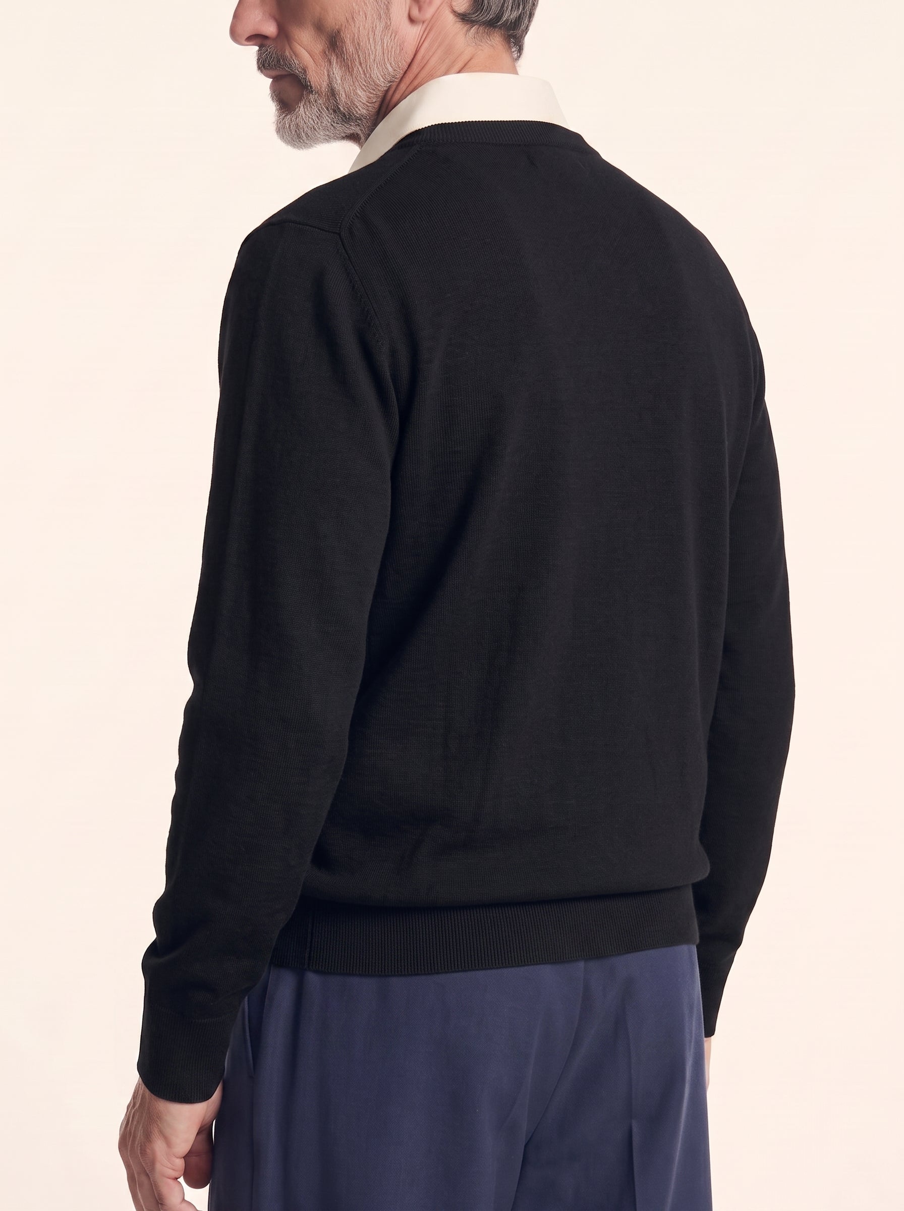 #Jet-Black_group A man shown from the back wearing a black long-sleeve knit sweater over a cream collared shirt and navy trousers, against a plain light beige studio background.