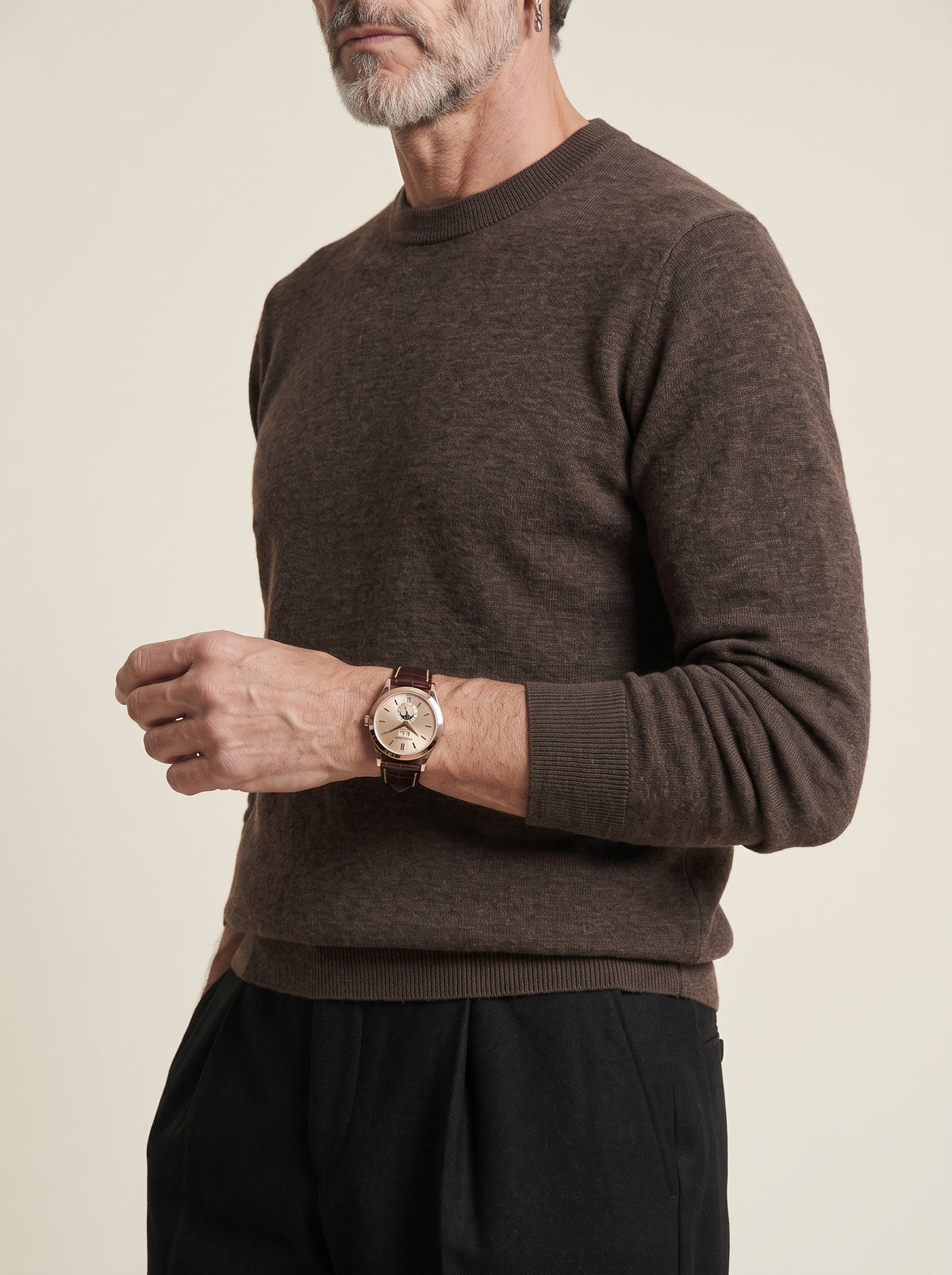 #Cedar-Brown_group A man wearing a dark brown long-sleeve knit sweater and black trousers, shown from the chest down against a plain light beige background. He is wearing a wristwatch on his left wrist.