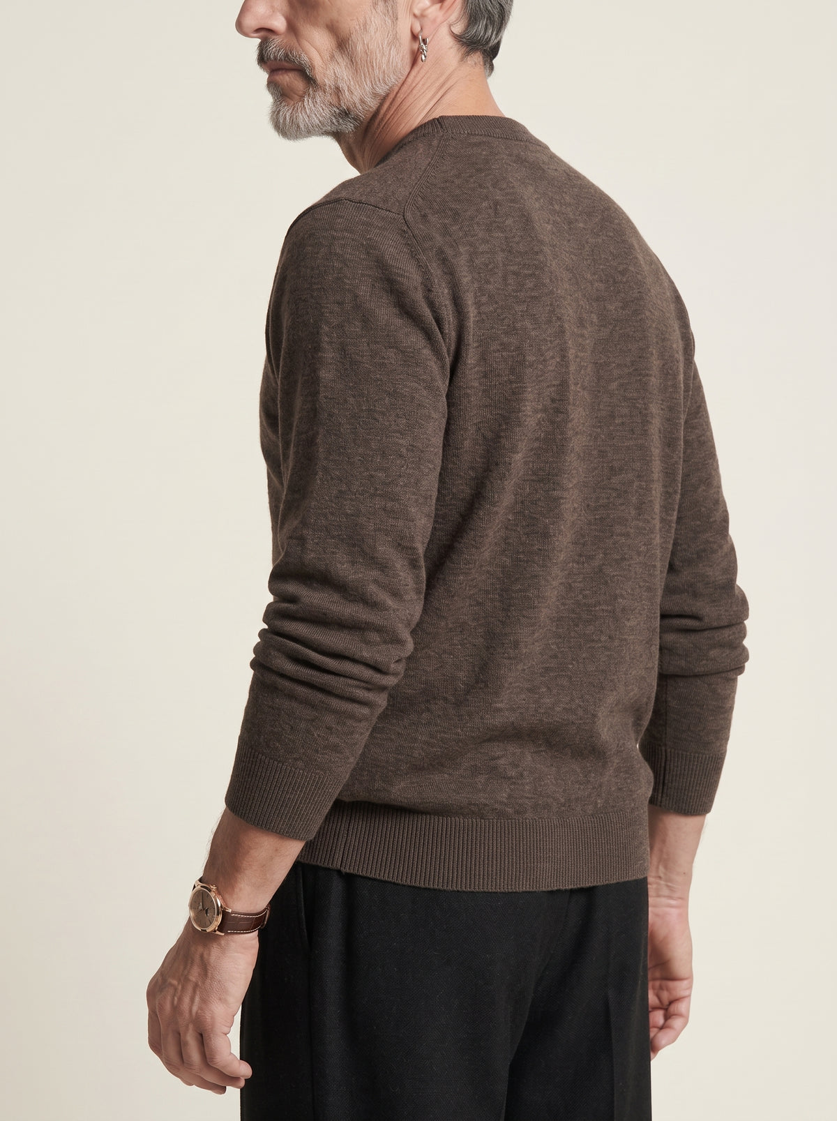 #Cedar-Brown_group A man wearing a brown long-sleeve knit sweater, shown from a side-back angle against a plain light beige studio background.