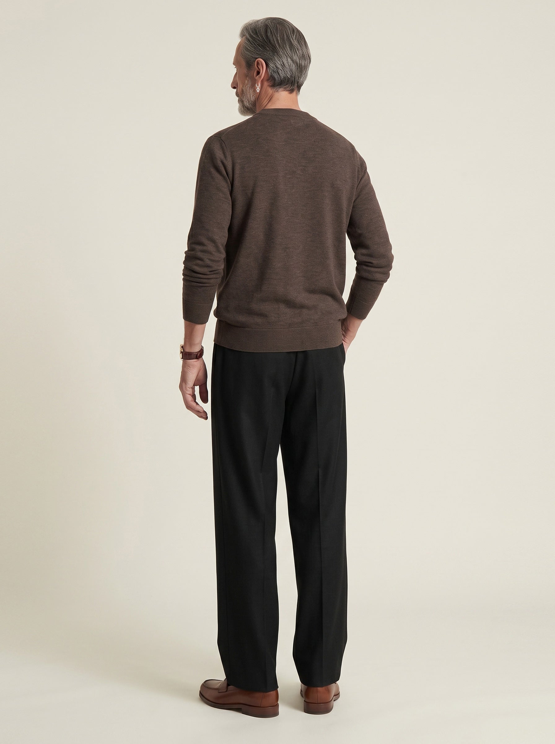 #Cedar-Brown_group A man viewed from the back wearing a brown knit sweater, black trousers, and brown leather shoes, standing against a plain light beige studio background.